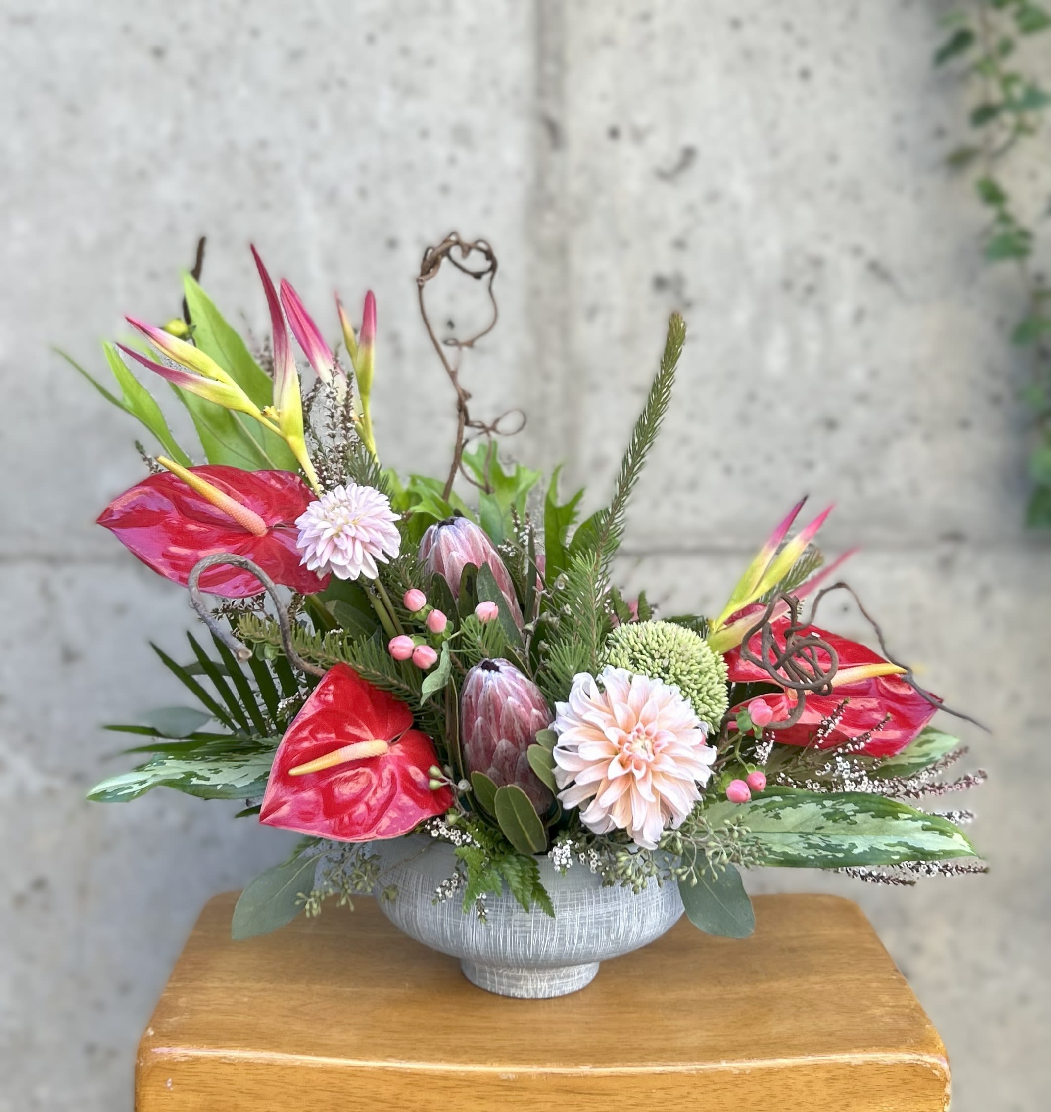 Tidalwave - Like the crest of a tropical sea, Tidalwave sweeps the eye with bold shapes and refreshing color. Striking pink anthuriums and proteas rise among soft blush dahlias, accented by cheerful yellow-and-pink heliconia and curling twigs for movement and drama. Textured greenery and berries add depth, while the arrangement’s low, ocean-toned vessel grounds it with coastal elegance. Perfect as a statement centerpiece, this design blends exotic vibrance with soft, romantic undertones — a floral tide you’ll never want to recede.