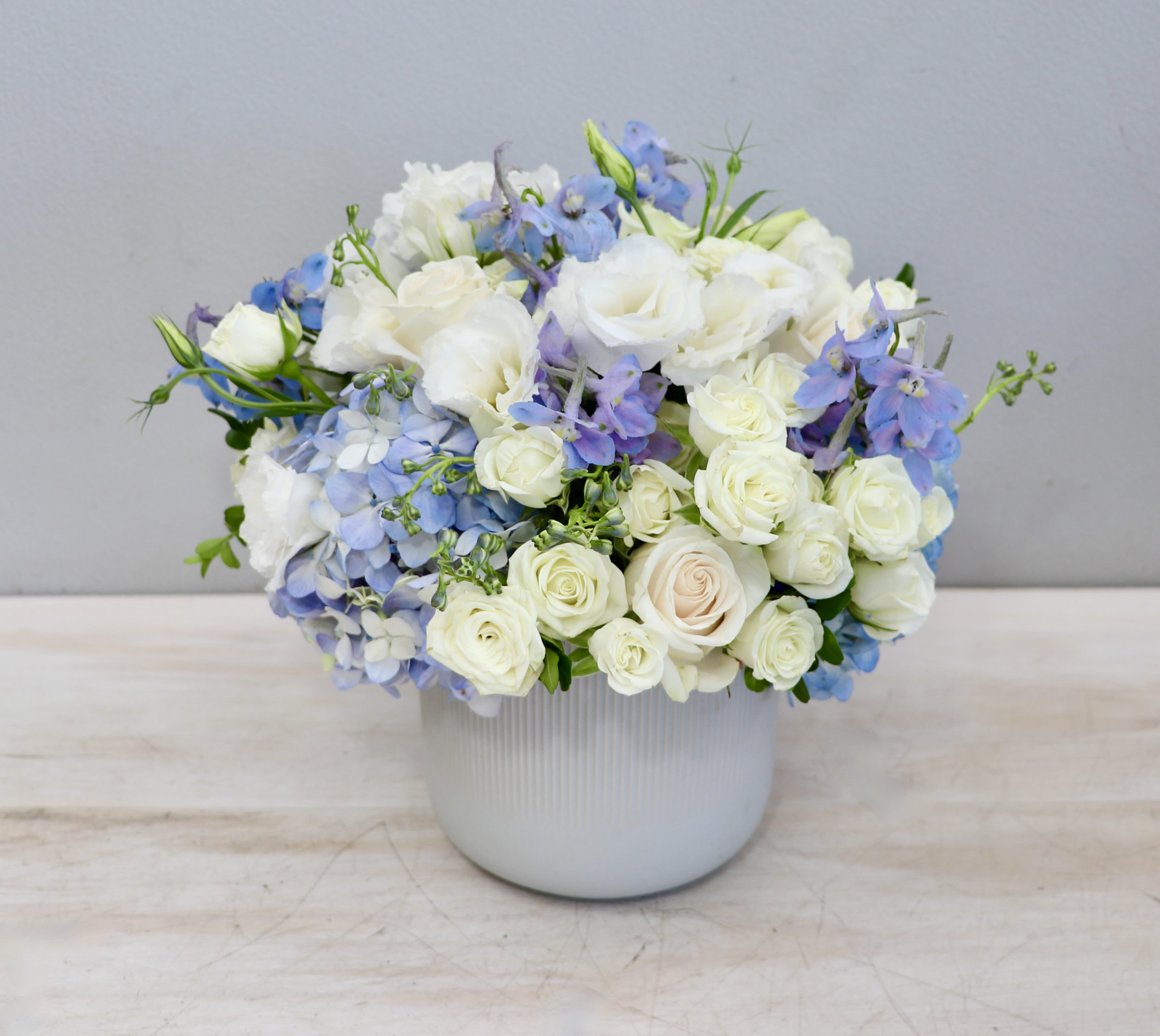 Blue Clouds - Weho Blue Clouds - Weho - This arrangement is made in a white ceramic vase with light blue delphinium and hydrangea flowers and white roses and lisianthus.