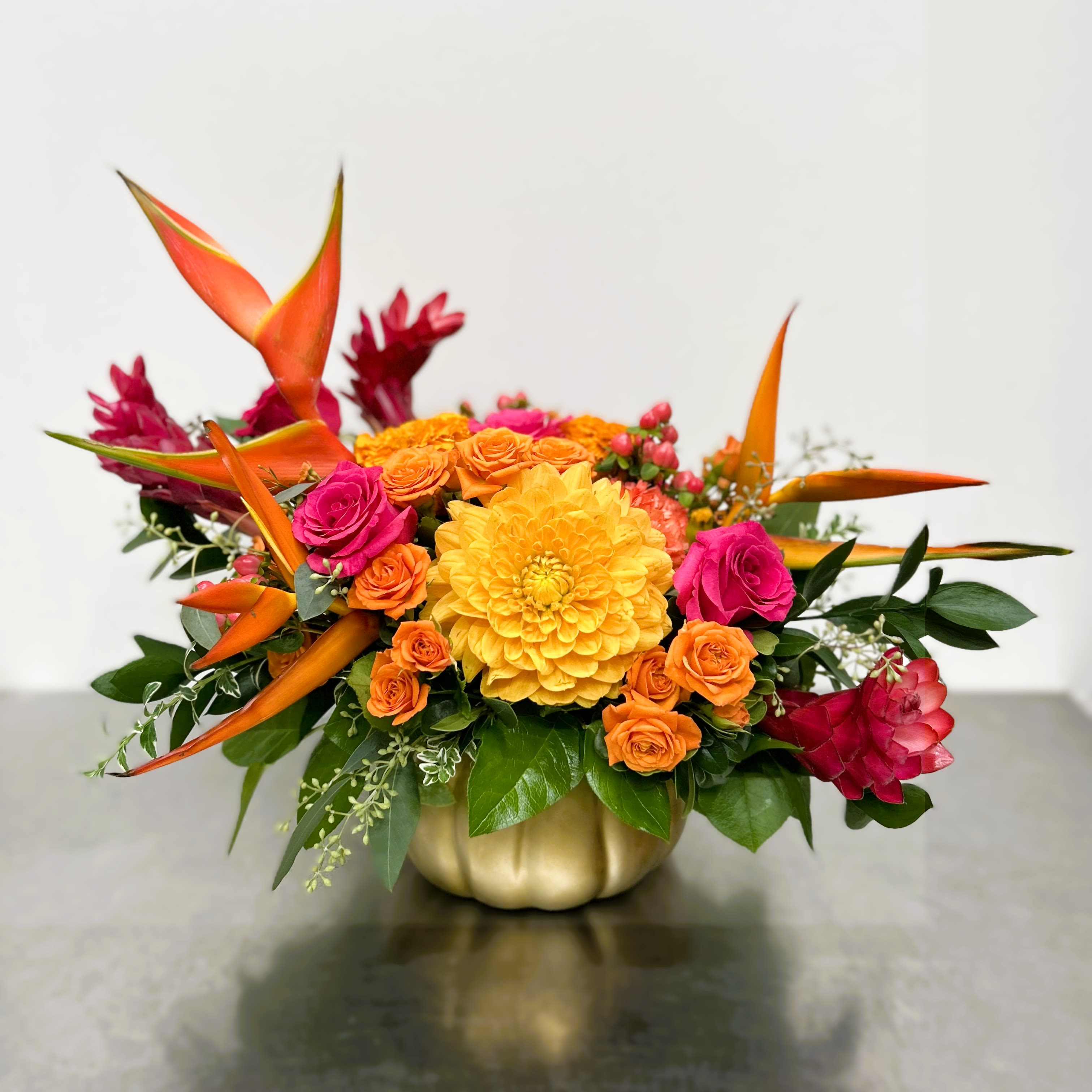 The Enchanted Gourd - Step into a fairytale with this radiant autumn treasure. Nestled in a shimmering gold pumpkin vessel, The Enchanted Gourd bursts with the tropical drama of heliconia, the exotic charm of pink ginger flowers, clusters of vibrant spray roses, and the sunny brilliance of golden dahlias. Lush greenery frames the blooms, creating a rich tapestry of color and texture that feels both magical and regal. Perfect for gifting, seasonal décor, or as the spellbinding centerpiece of your next gathering.