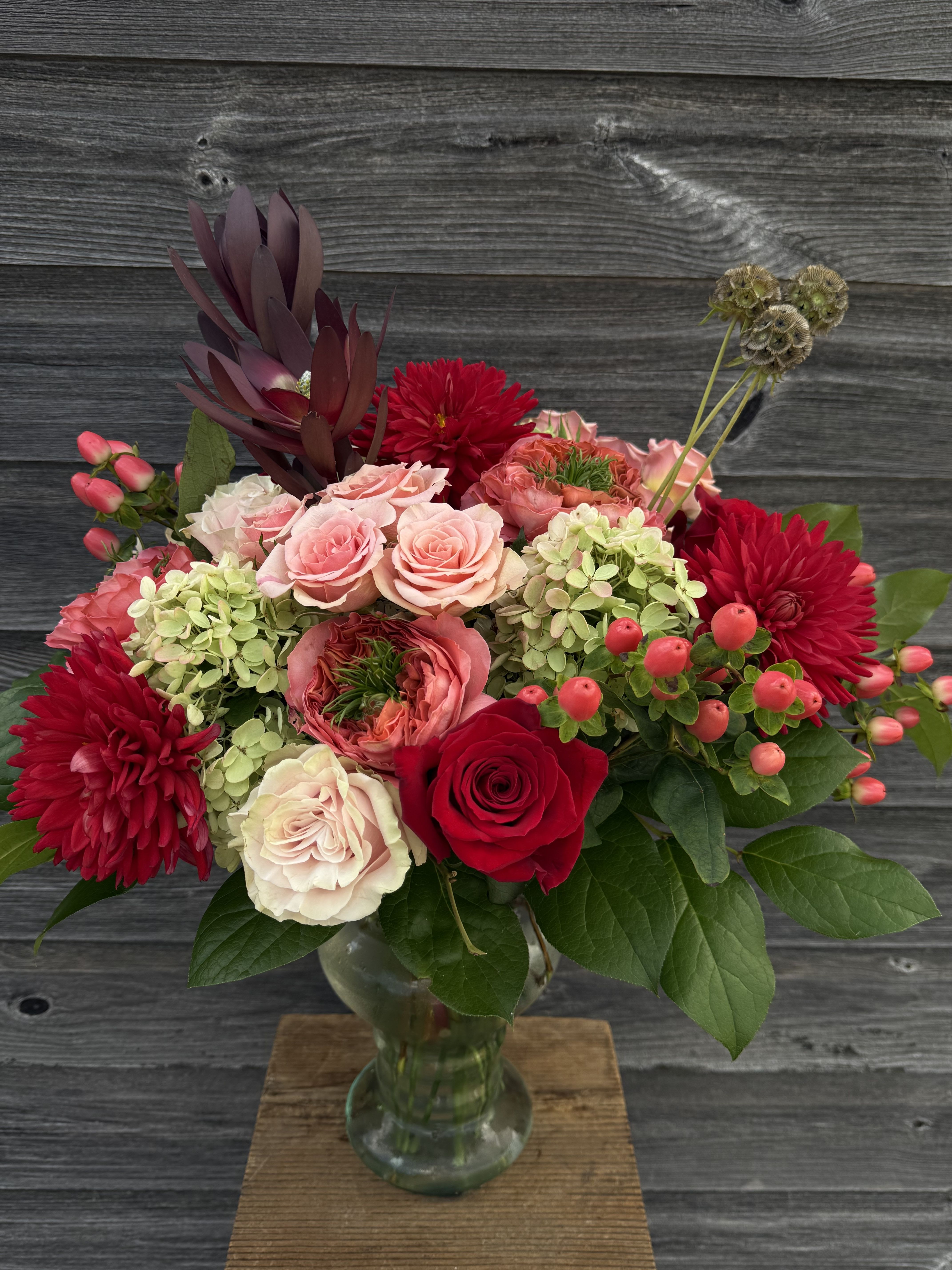 Elegant Red and Coral (Limited Edition) - Medium height glass pedestal vase containing dahlias, hydrangea, roses, spray roses, hypericum berries, protea and star scabious .
