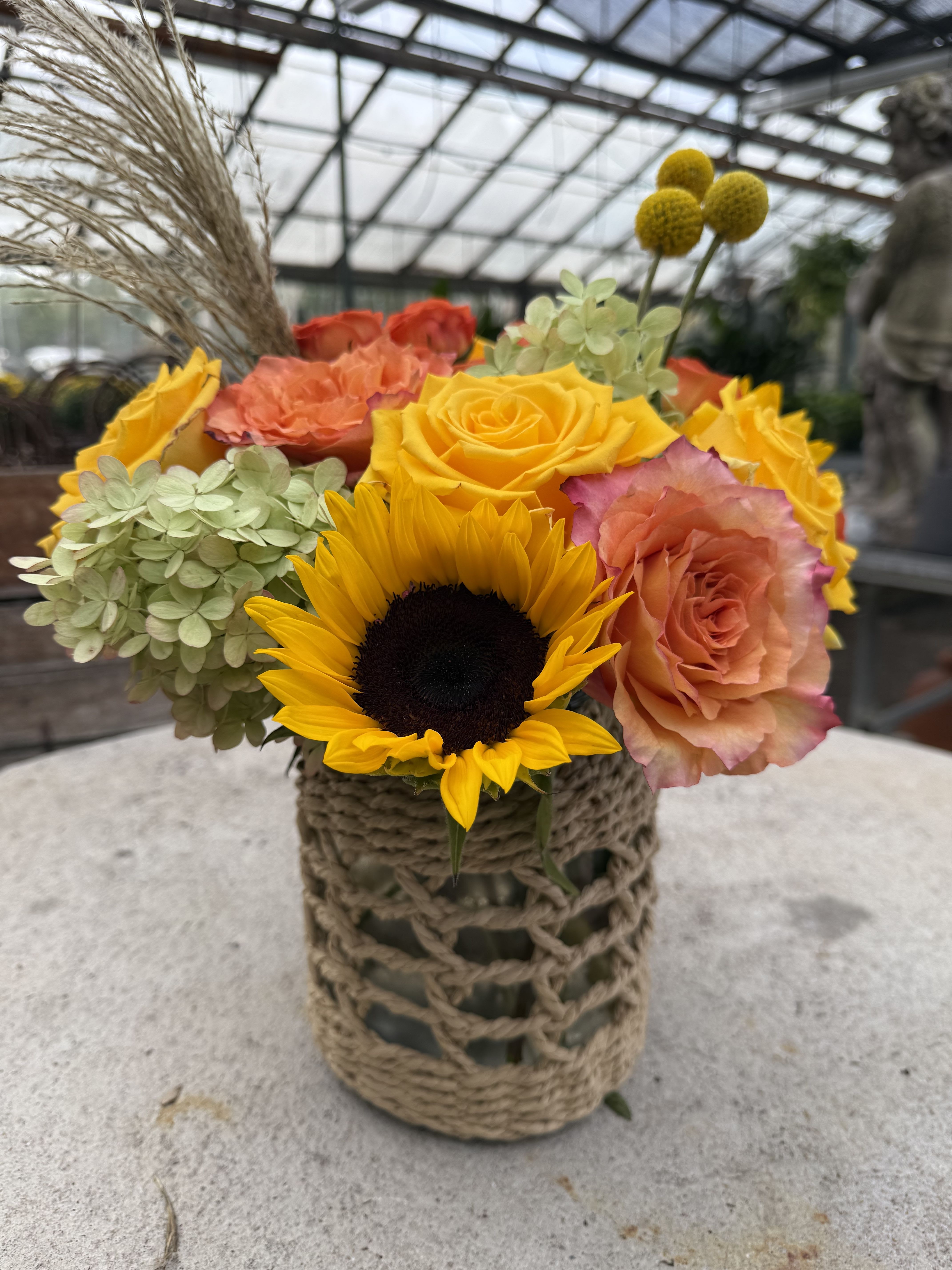 Whimsical Fall Harvest -- Limited Edition - Bright yellow and orange flowers, including sunflowers, billy balls, roses, spray roses and chartreuse hydrangea adorn a natural jute covered vase. 
