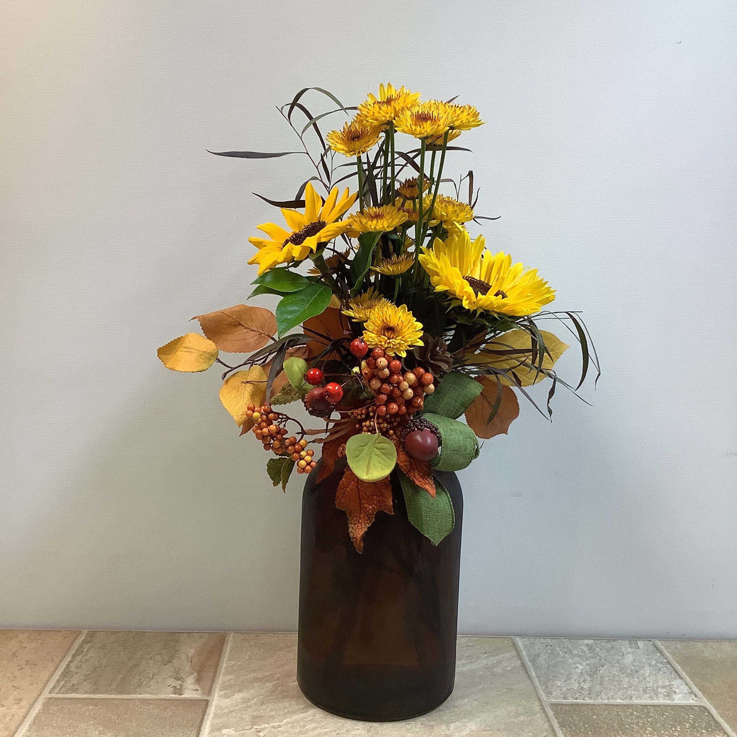Amber Fields Amber Fields - This is a tall, rustic fall-inspired bouquet arranged in a deep amber glass vase. The centerpiece features bold golden-yellow sunflowers and smaller yellow daisy mums, accented with airy brown grasses for height and texture. Tucked among the blooms are pinecones, faux berries, and autumn leaves, adding warmth and seasonal charm. A touch of green ribbon softens the arrangement while complementing the natural earthy palette. It has a cheerful yet cozy harvest feel, perfect for autumn décor or as a seasonal gift.