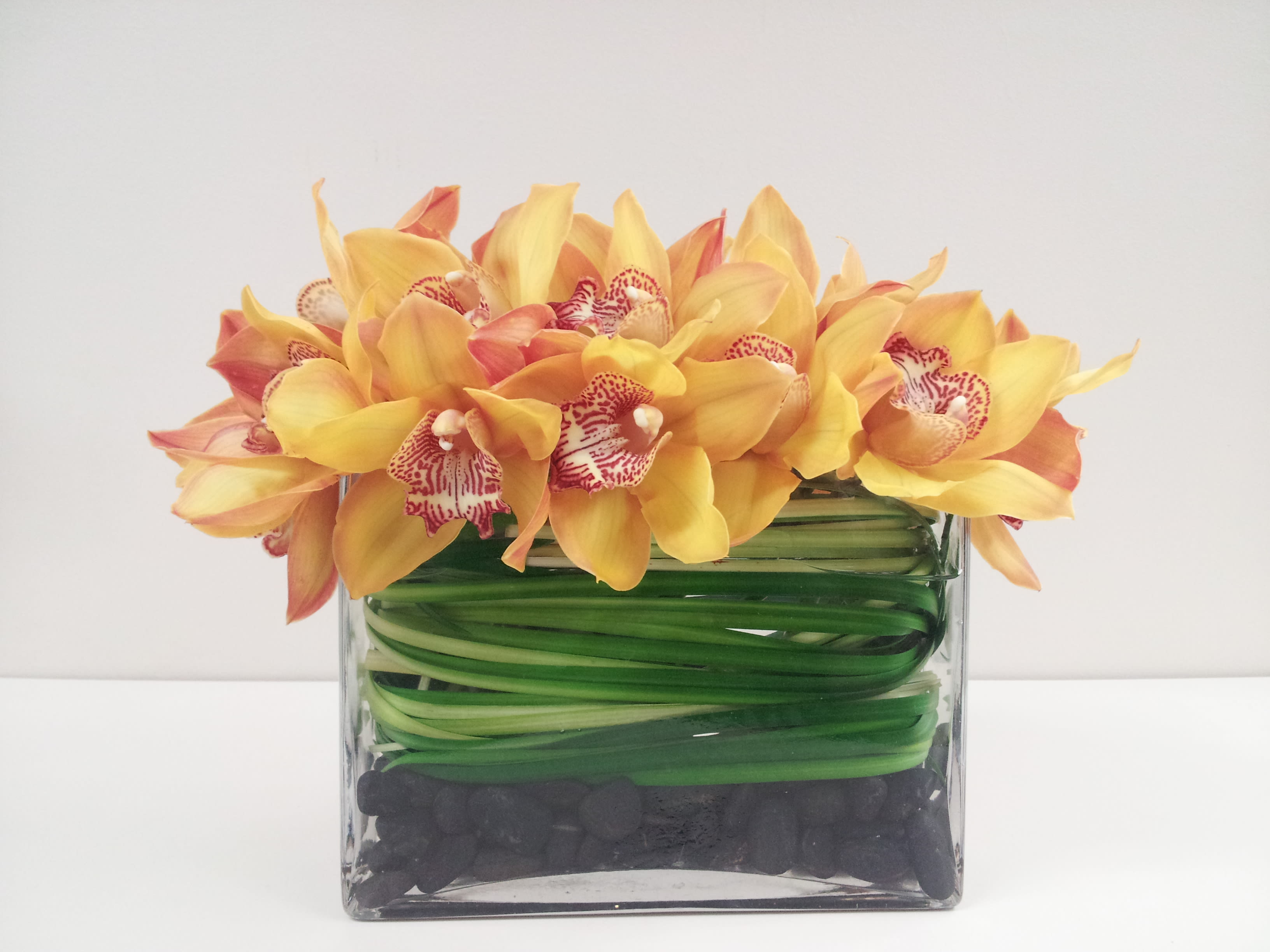 Hawaiian Sunset - Beautiful sunset colored cymbidium orchids nestled in sleek black river rocks in a short rectangular grass wrapped vase.