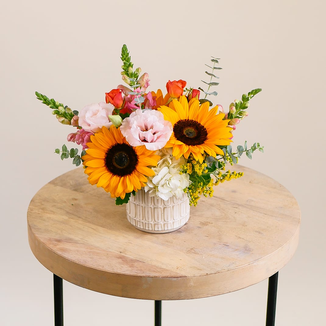 Sunbeam Delight - Add warmth and sunshine to any space with Sunbeam Delight, a cheerful sunflower arrangement designed to brighten someone’s day. This fresh flower arrangement features vibrant sunflowers, soft pink lisianthus, white hydrangea, orange spray roses, snapdragons, solidaster, and accents of eucalyptus—artfully arranged in a textured ceramic vase. Perfect as a birthday gift, thank-you flowers, or a summer centerpiece, this sunflower floral arrangement is a stunning choice for home decor or special celebrations. Bring seasonal joy and natural beauty to your loved ones with this radiant, handcrafted design. Arrangement approximately measures 10&quot; tall and wide with flowers.
