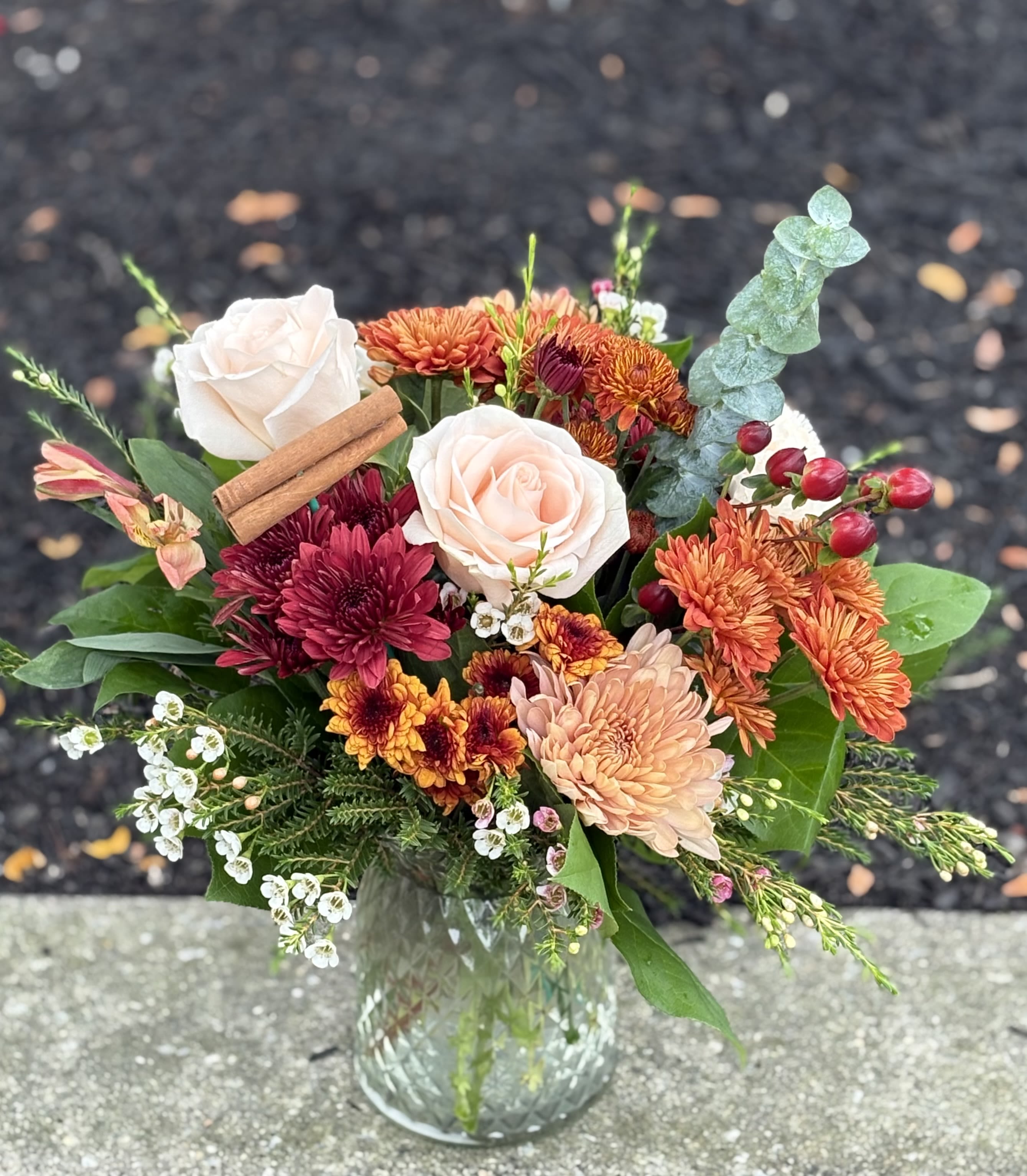 Cinnamon Spice - Cozy colors and cinnamon sticks make this arrangement a great choice for the fall season!!! Seasonal poms, berries, roses, fillers and greenery in a clear glass “quilted” vase. Arrangement is approximately 14” x 14”, making it perfect for a kitchen table or island, entry or accent table. Upgraded versions include a few more stems for extra fullness.