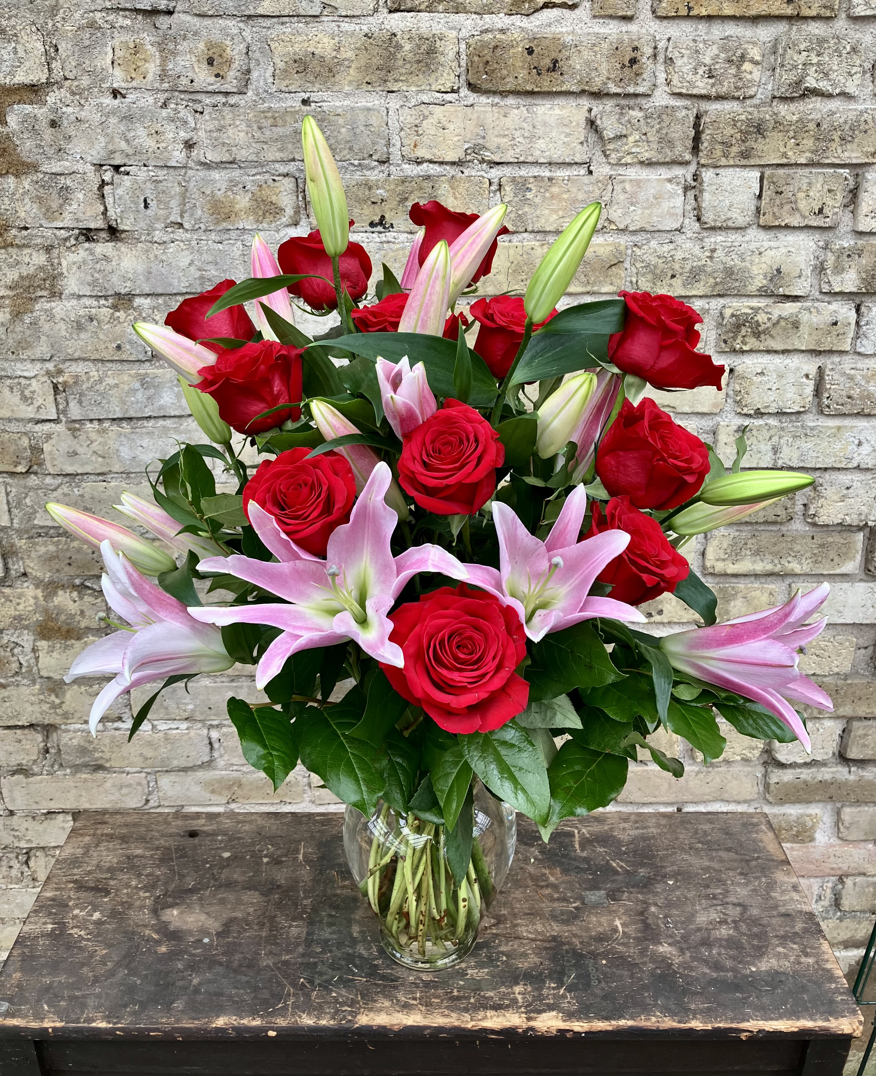 Kiss Me Kiss Me - One Dozen Red Roses Arranged in a Clear Glass Vase With Greens and Lilies. (Color of lilies may vary depending on availability)
