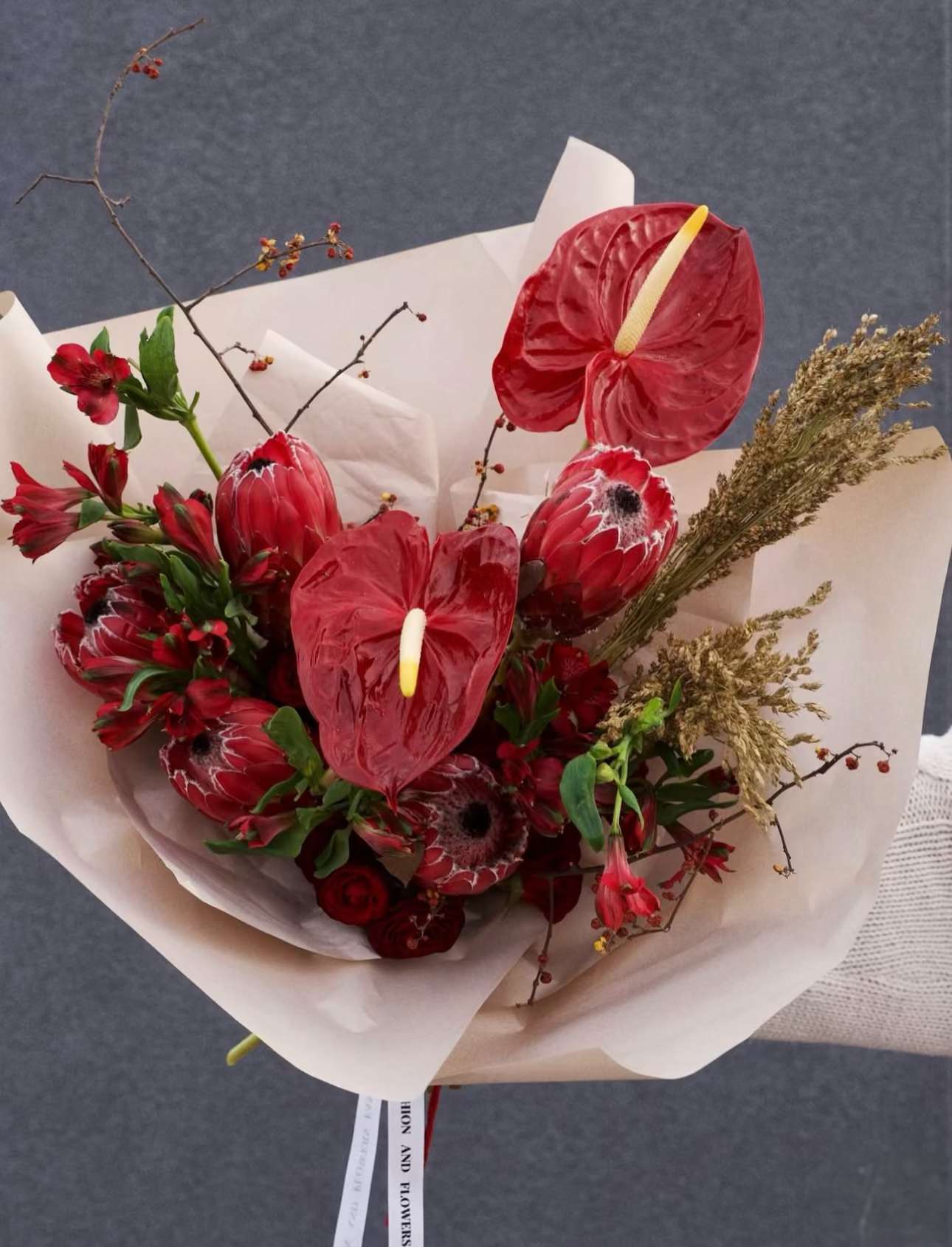 Pomegranate Elegy - A sophisticated bouquet in deep crimson and pomegranate shades. Featuring bold anthuriums, exotic proteas, and delicate seasonal accents, it embodies elegance, strength, and timeless style.