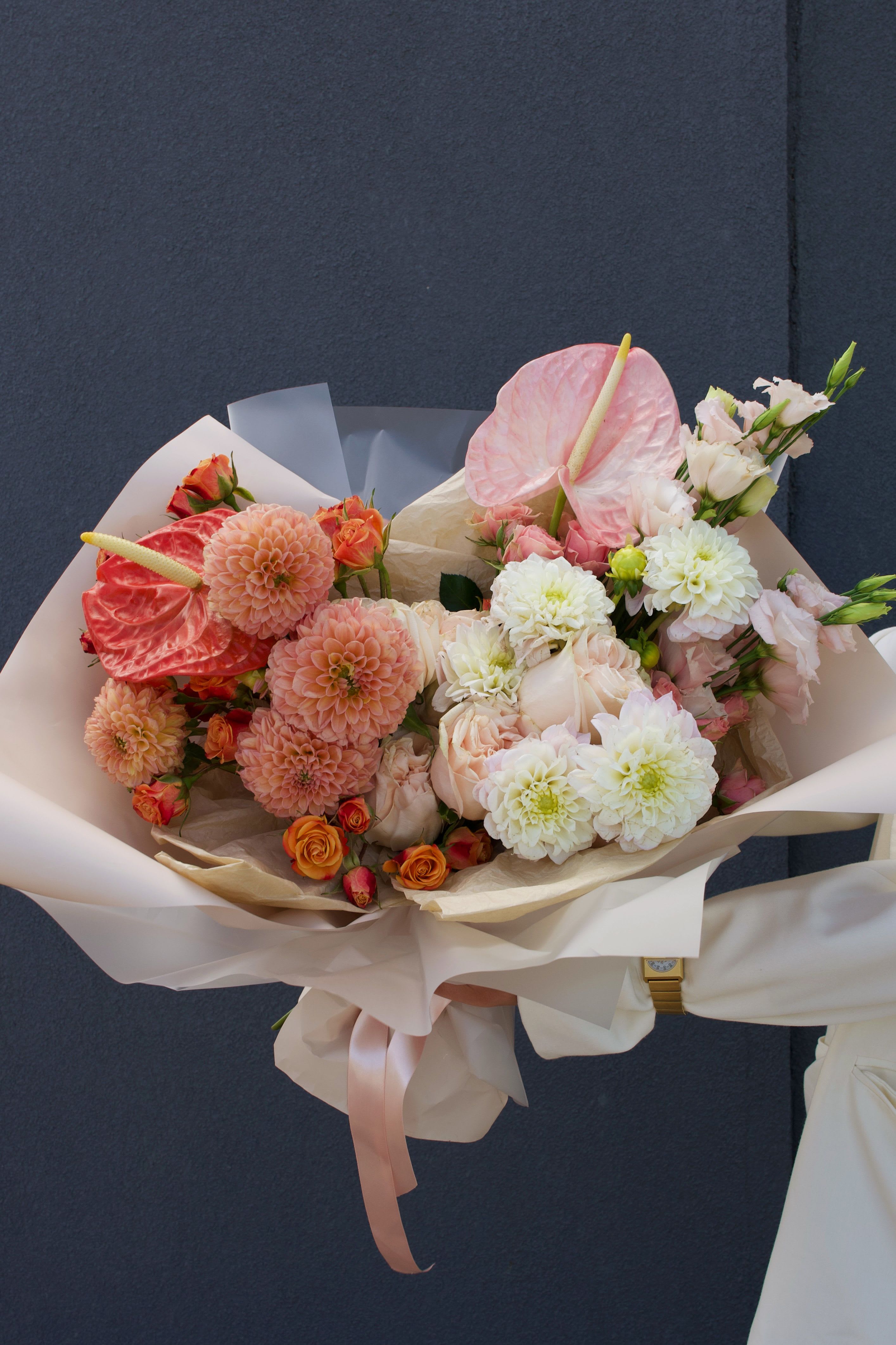 Powder Blush - A modern statement bouquet where soft blush meets coral accents. Anthuriums, dahlias, and garden roses come together in an elegant harmony of color and texture.