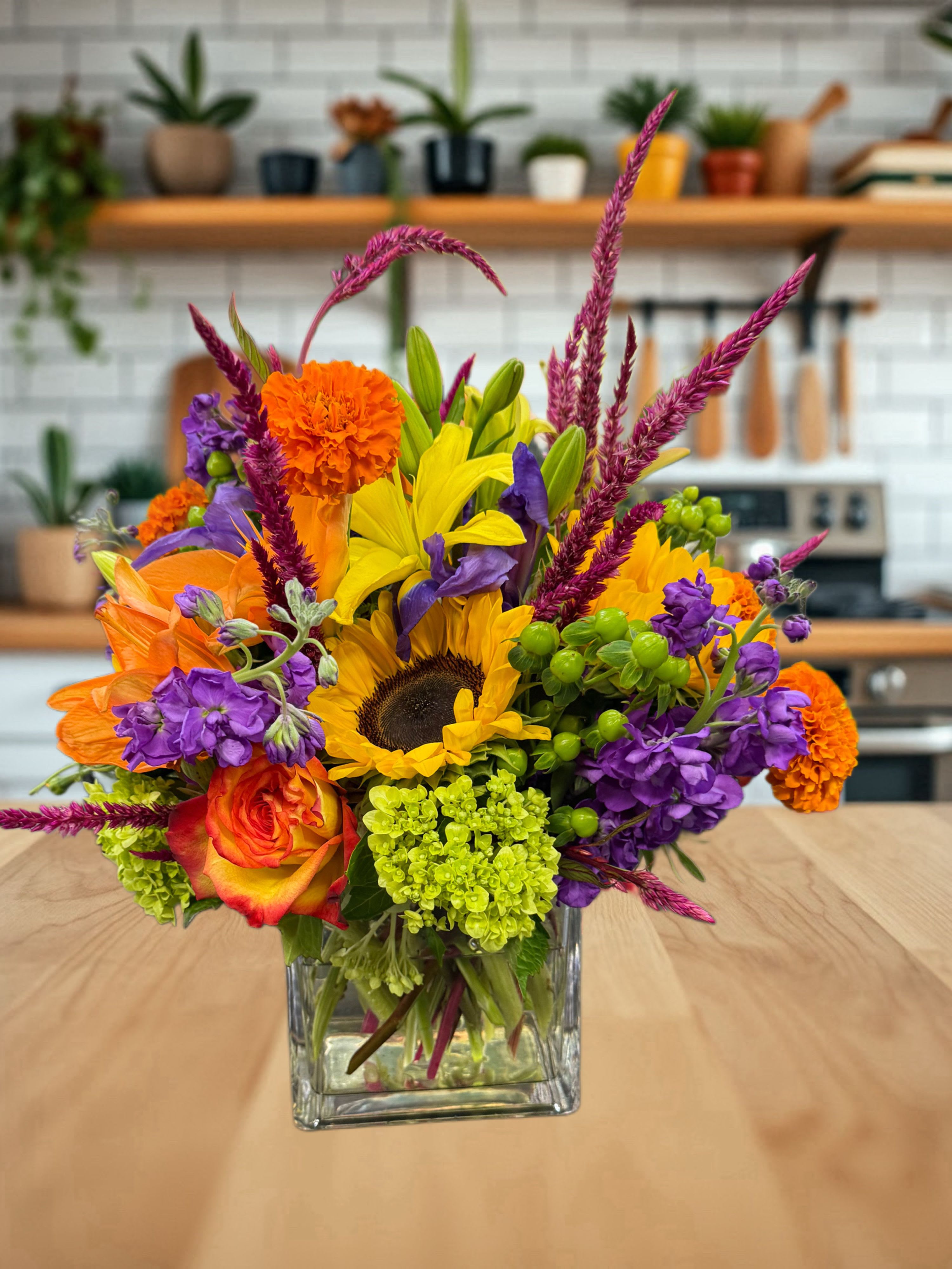 Autumn vibes  - This vibrant cube vase arrangement bursts with bold, cheerful colors and a mix of textures. At the heart, a bright sunflower is surrounded by orange roses, fiery carnations, purple stock, and delicate iris blooms. Accents of green hydrangea and hypericum berries add freshness, while magenta celosia spikes bring height and drama. Arranged in a clear glass cube, this design combines warmth, playfulness, and elegance—perfect for brightening any room or celebrating a joyful occasion.
