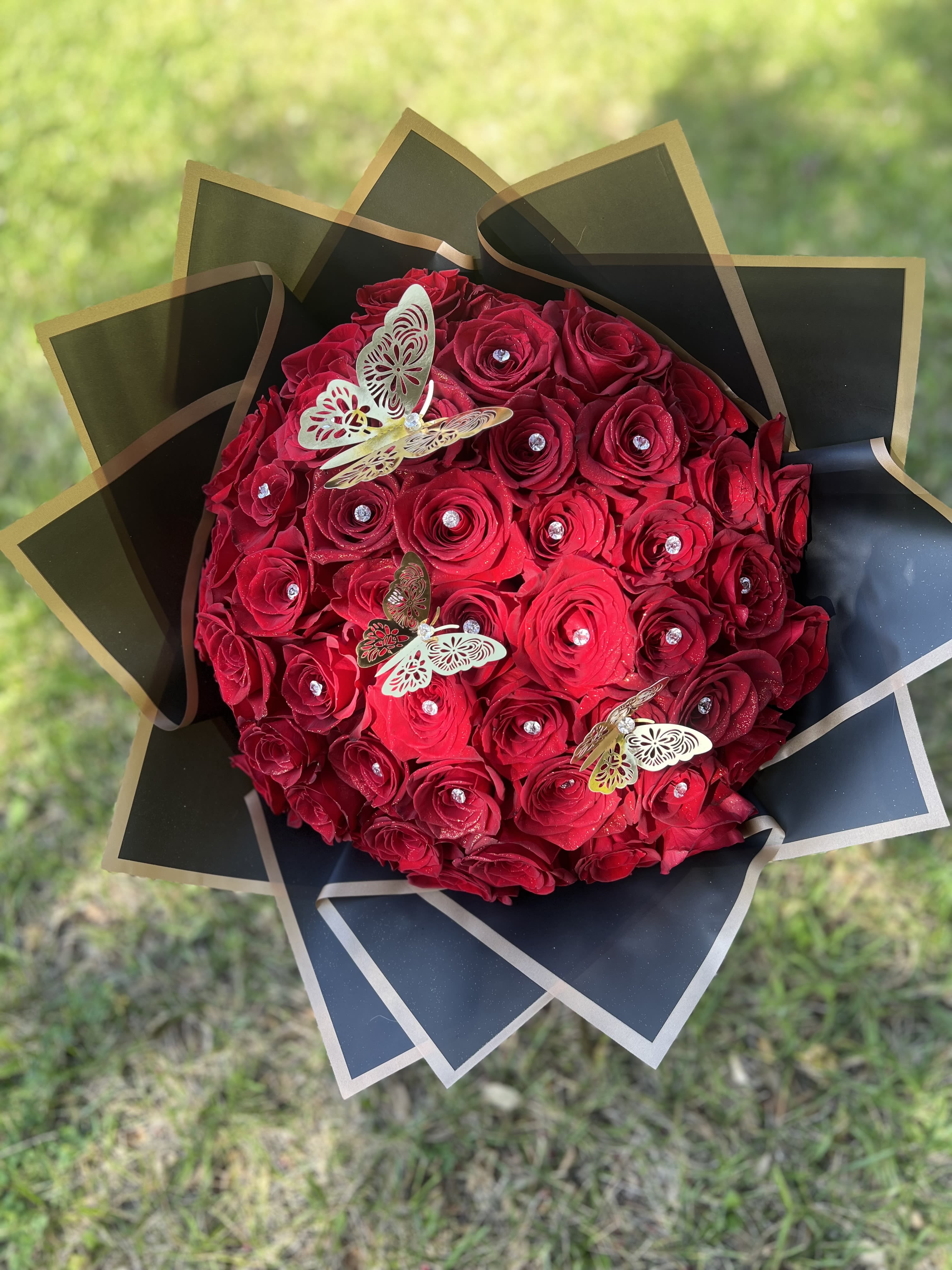 Red Roses Bouquet Red Roses Bouquet - A breathtaking arrangement of radiant red roses, each bloom adorned with a sparkling crystal accent for a touch of timeless luxury. Nestled within layers of sleek black and gold wrapping, the bouquet is graced with delicate golden butterflies, symbolizing transformation, love, and beauty. Perfect for anniversaries, romantic gestures, or unforgettable celebrations, this bouquet captures passion and sophistication in one unforgettable gift.