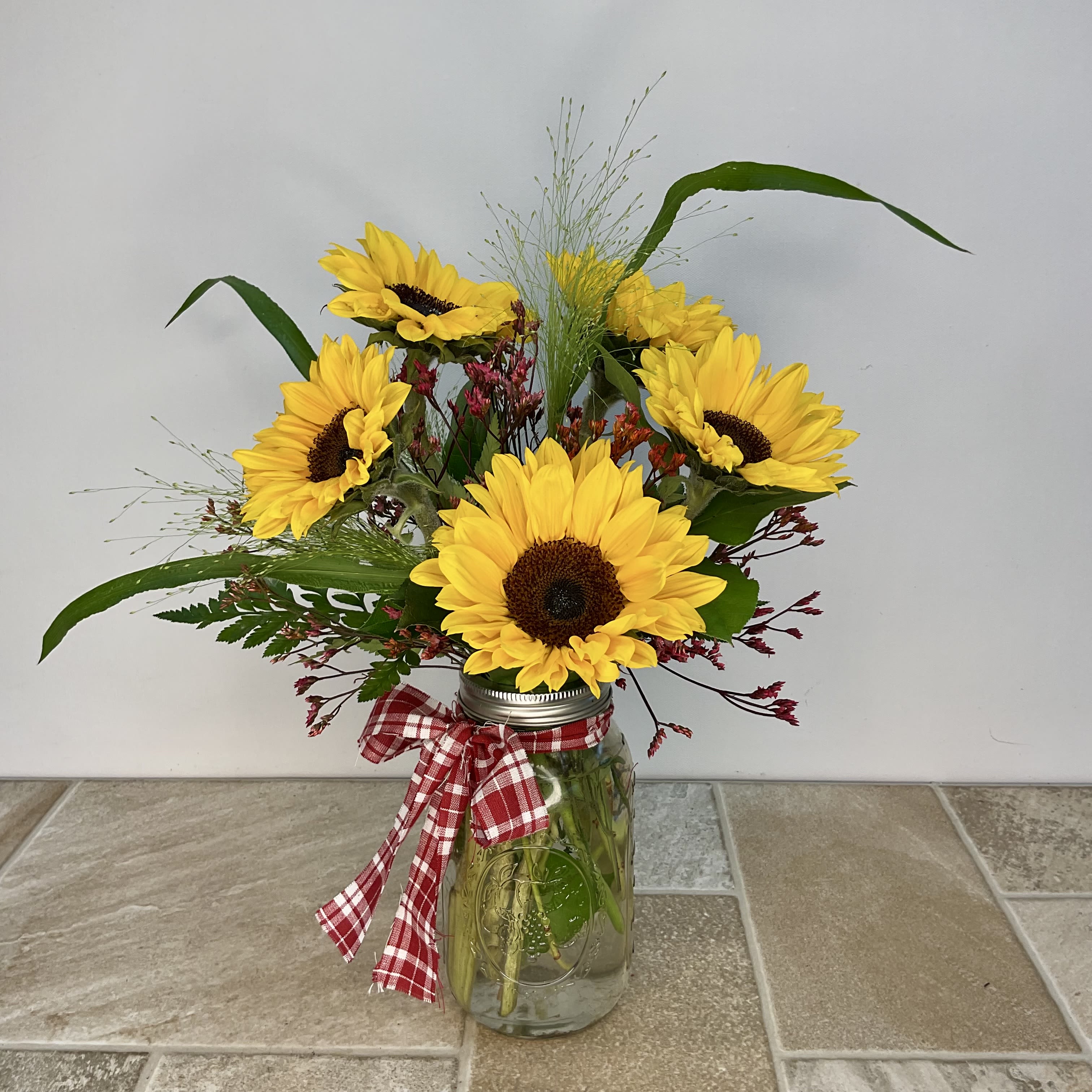 Country Sunshine Country Sunshine - Bright yellow sunflowers radiate sunshine and country charm with their bold, brown centers and golden petals. Delicate red & orange accent flowers are interspersed throughout, adding pops of color, and grasses and leafy greens give the arrangement a natural, just-picked-from-the-meadow feel. A red and white plaid ribbon tied around the mason jar full of blooms enhances the country-style presentation and adds a playful, nostalgic touch. Approximate size: 12" w x 16" h Primary flower: Sunflower