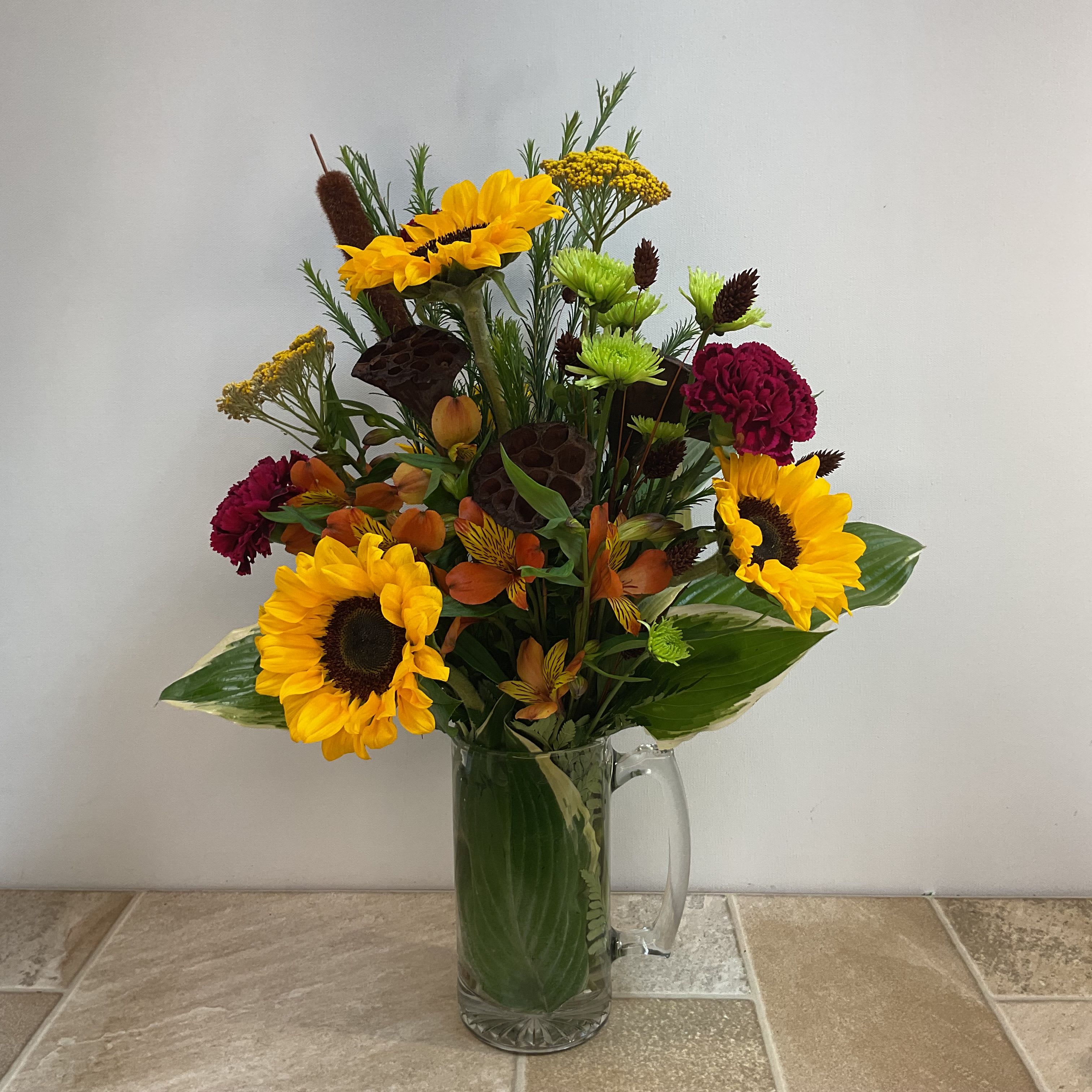 Rustic Meadow Rustic Meadow - This outdoorsy floral arrangement evokes the feel of a late-summer hike through sunlit fields and woodlands. Bold golden sunflowers serve as the focal point, radiating warmth and strength. Burgundy carnations and orange alstroemeria add rich, earthy tones, while bright green athos button poms and yellow yarrow bring a touch of natural vibrance. Textural elements like dried lotus pods and a cattail give a rugged, woodland edge. Lush variegated hosta leaves and sturdy greenery anchor the piece in a large clear glass mug wrapped inside with a large hosta leaf—adding to its natural, utilitarian charm. This arrangement is both grounded and vibrant, perfect for those who appreciate the wild, unrefined beauty of the outdoors. Approximately 16" wide x 22" high Primary elements: sunflower, carnation, alstroemeria, pom, yarrow, lotus pod