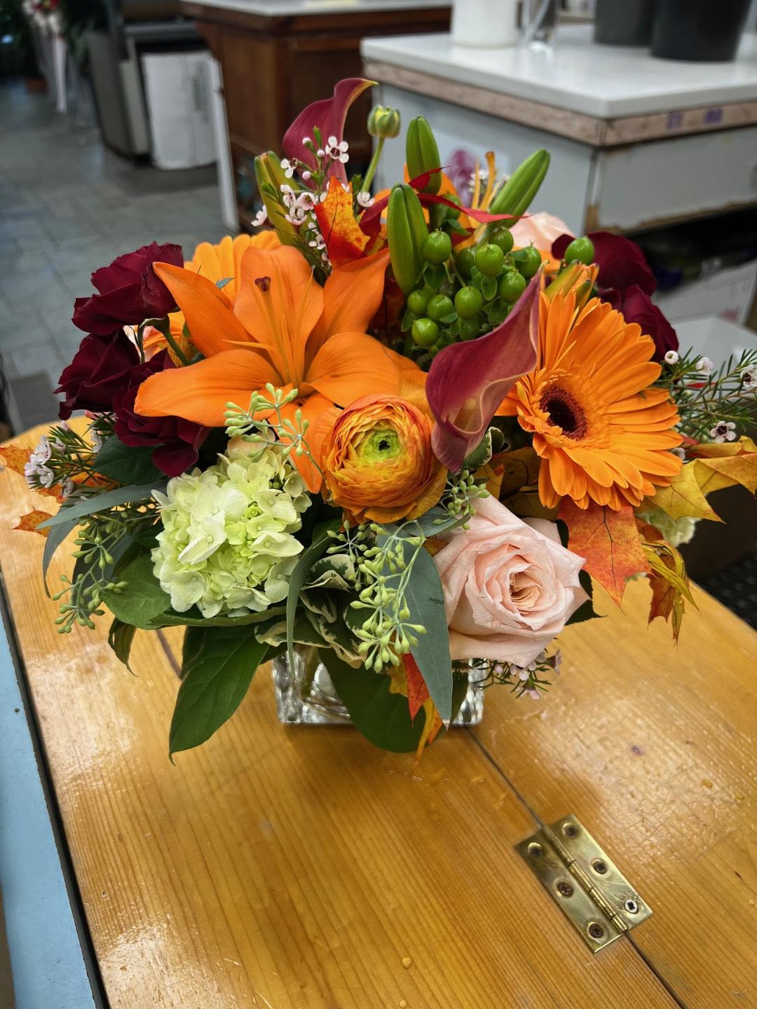 SEPTEMBER CENTERPIECE - MIXED FALL CENTERPIECE OF ROSES, SPRAY ROSES, LILIES, HYDRANGEAS, GERBERA DAISIES, RANUNCULUS, MINI CALLA LILIES, HYPERICUM BERRIES, WAXFLOWER, SEEDED EUCALYPTUS, AND FALL LEAVES ARRANGED ALL-AROUND IN A CLEAR GLASS CUBE 
