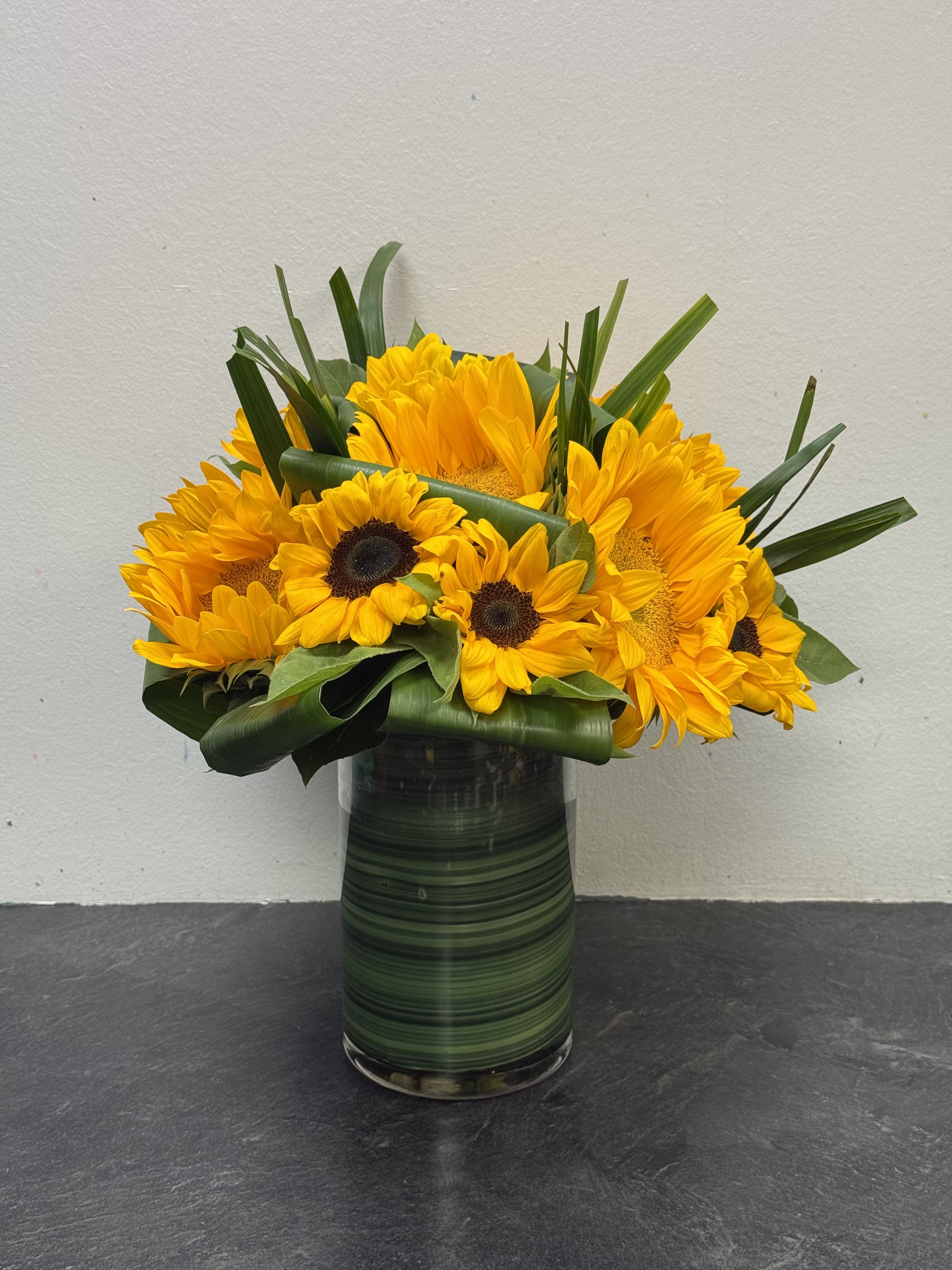 Golden Radiance Vase - A bold and cheerful display of radiant sunflowers, artfully arranged in a clear cylinder vase wrapped with lush green foliage. This vibrant design captures the warmth of sunshine and the joy of summer, making it a perfect gift to brighten any home, celebrate milestones, or send a heartfelt smile.