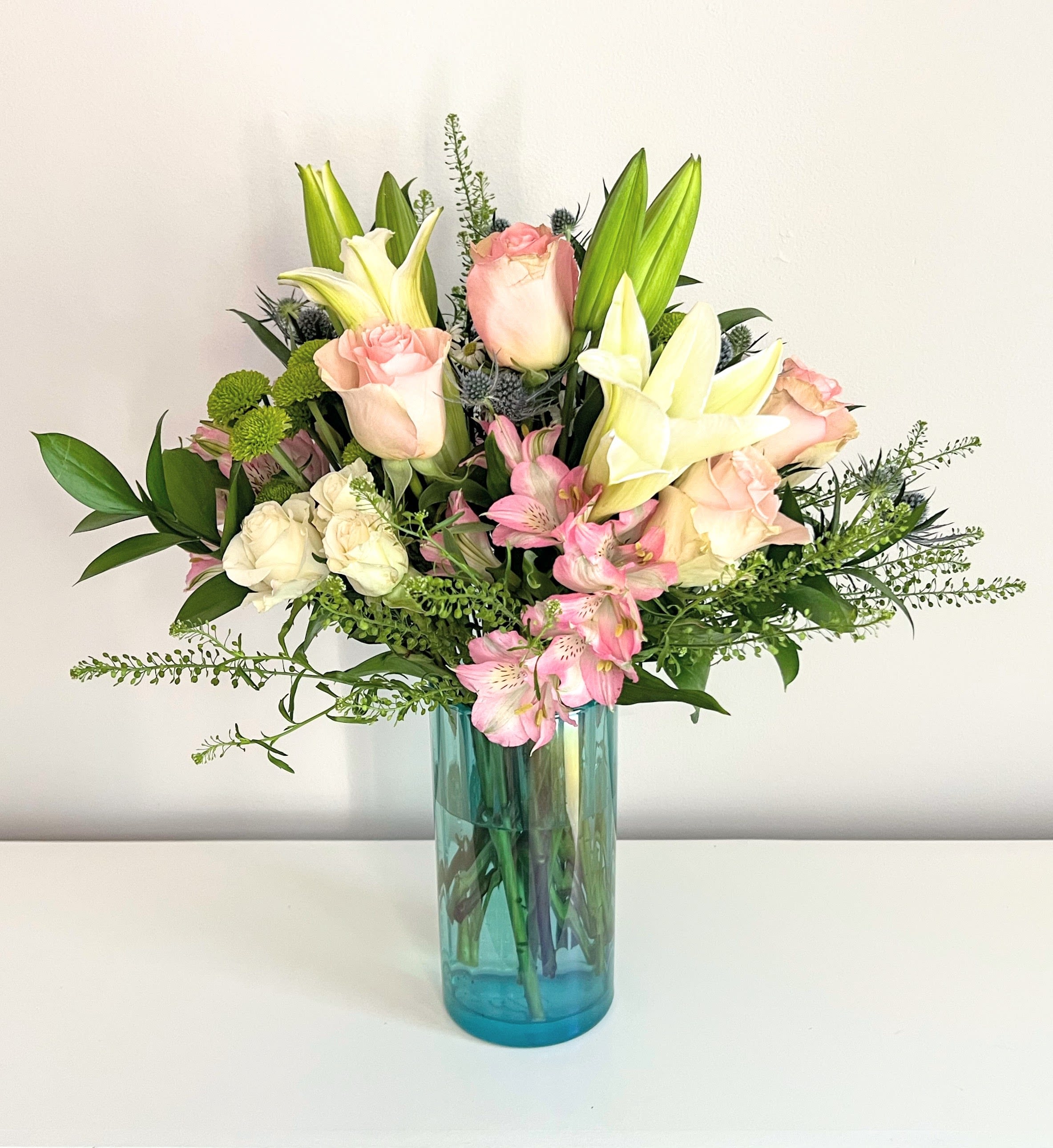 Blushed Beauty - A lush floral arrangement full of fragrant white lilies, light pink roses, light pink alstroemeria, spray roses, green button poms &amp; thistle accented with greenery. Arranged tall in a beautiful premium aqua blue glass vase. Perfect for all occasions.   Measures approx. 18&quot; tall and 16&quot; wide Orientation: All around  