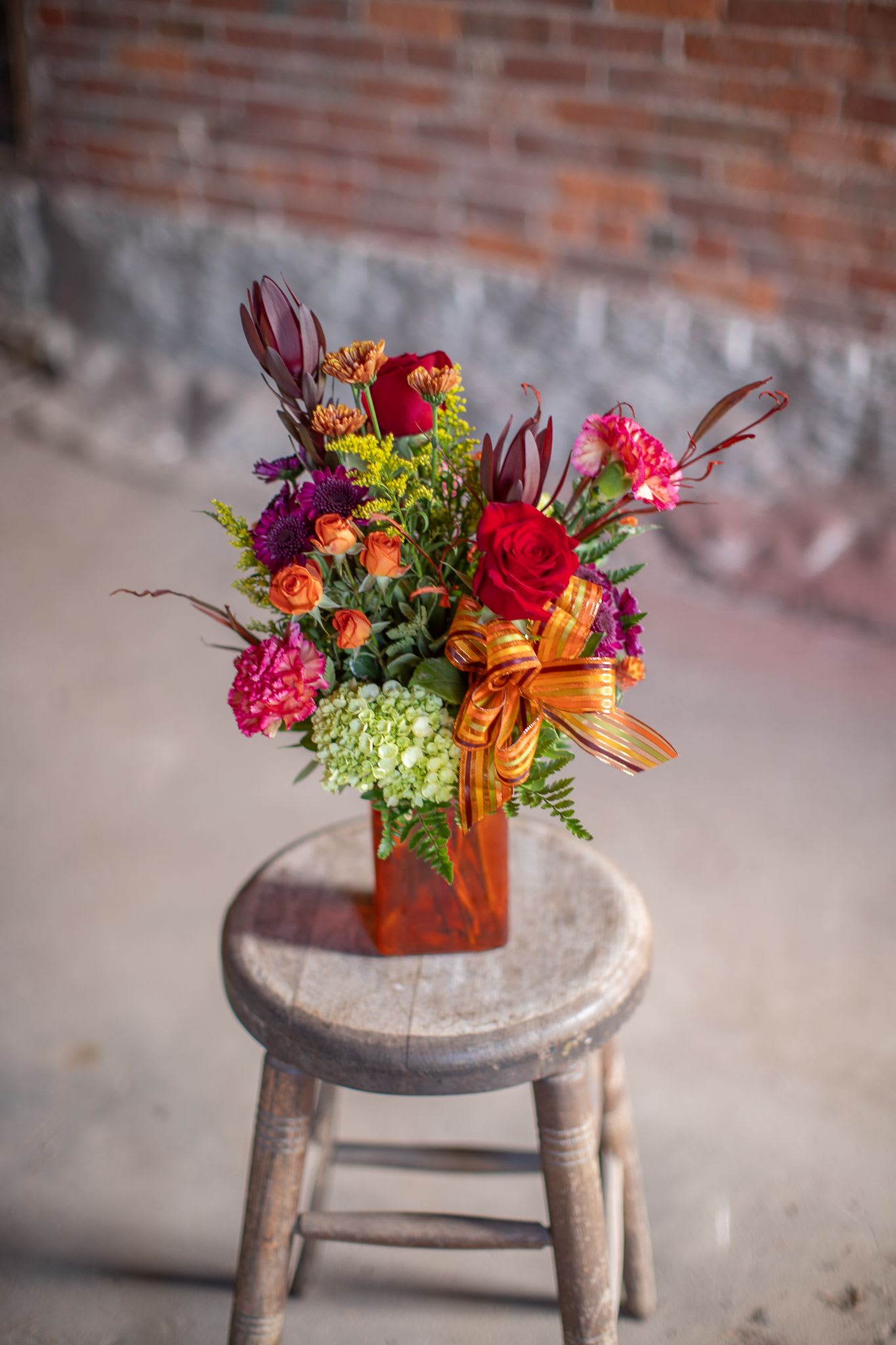 Fall Flame - A rectangular shaped vase in an autumnal hue (orange, green, rust, or red) is fill with delightful rich seasonal flowers. Complete with a festive striped satin and sheer bow. This design features mini green hydrangea, roses, spray roses and bright orange grevillea which mimics the look of flames at your autumn bonfire. Dianthus and seasonal pom pons complete the design. Shades and varieties by vary from photos but will be autumnal in essence. 