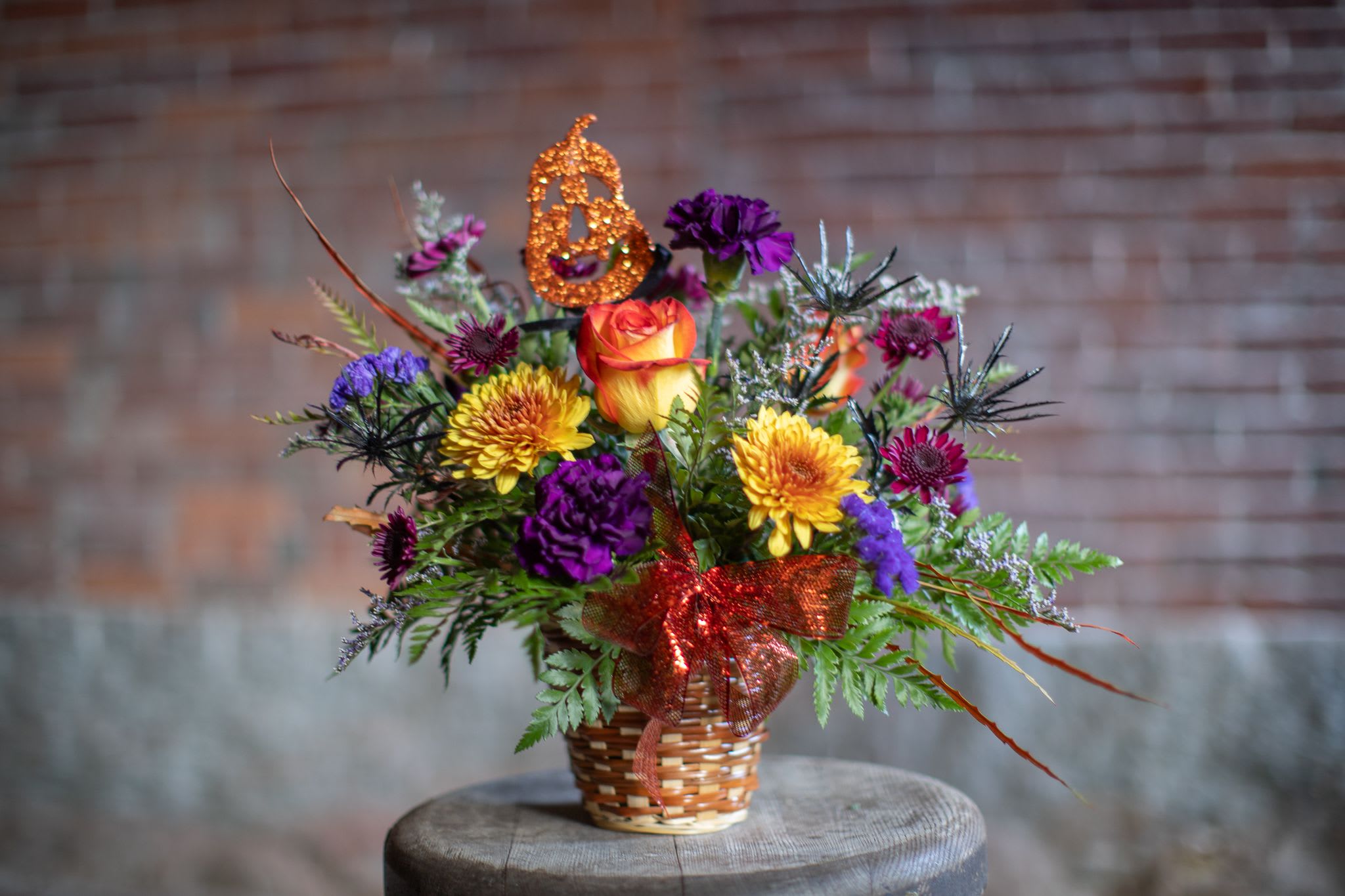 Oh My Gourd! - Lydia's fun Halloween inspired design. A sparkling Jack-O-Lantern pick is brought to life with festive roses, deep purple &quot;Moon Vista,&quot; carnations, assorted fall pom pons AKA spray chrysanthemums, black thistle adds depth and created the look of spooky spiders creeping around the floral arrangement. 