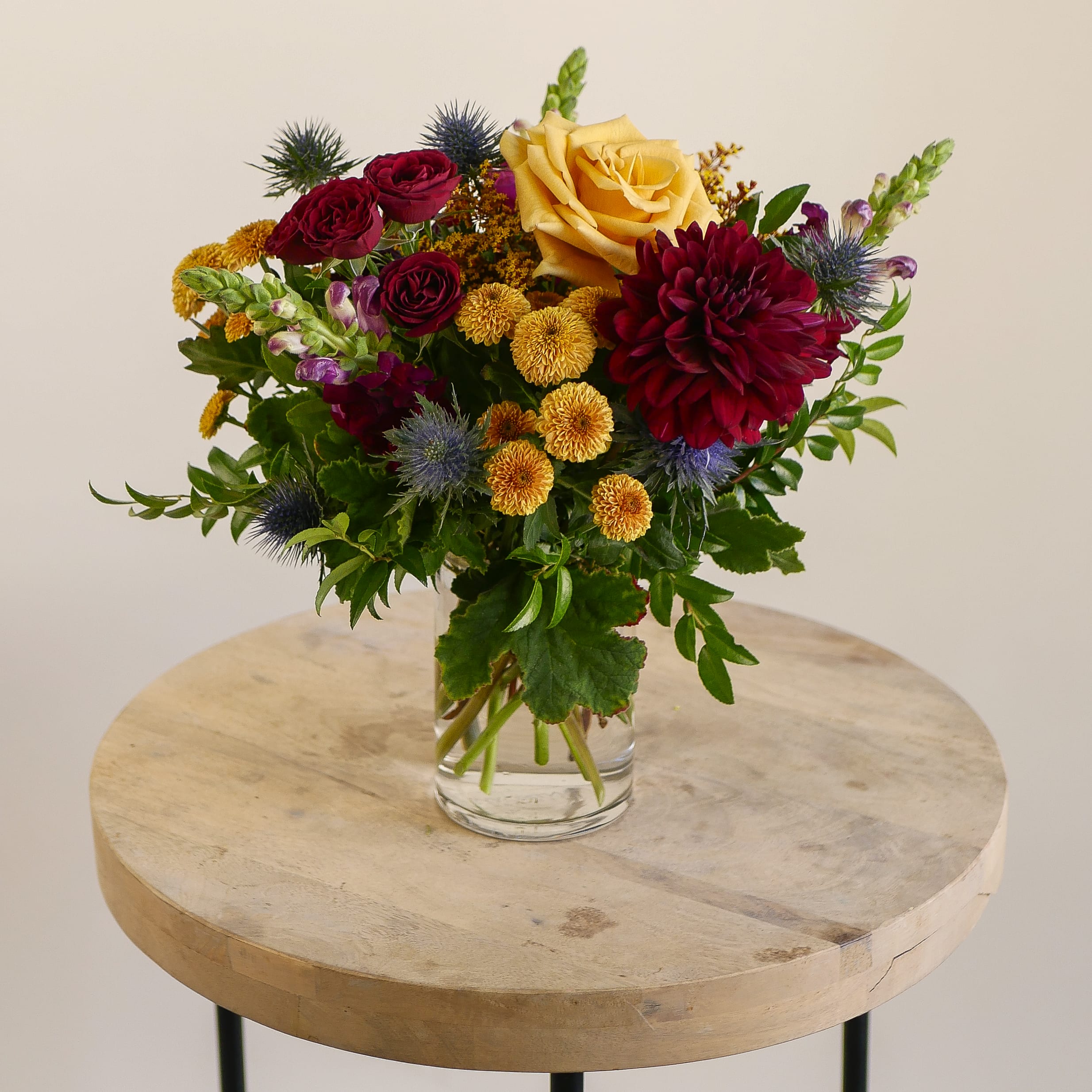 Dahlia Dusk - Make a bold seasonal statement with our Dahlia Dusk arrangement, a lush design featuring rich jewel tones and golden accents. This arrangement highlights a striking burgundy dahlia paired with deep red spray roses, a golden garden rose, vibrant mums, and unique blue thistle for a pop of texture. Accented with fresh greenery and styled in a clear glass vase, it’s both elegant and dramatic.  Perfect for fall gatherings, birthdays, or as a stylish gift, this dahlia arrangement brings warmth and sophistication to any space. Handcrafted by our expert florists, it’s a unique centerpiece that celebrates the beauty of autumn with rich colors and fresh seasonal blooms.  Available for local West Los Angeles flower delivery — surprise someone special with the vibrant charm of dahlias and roses this season.