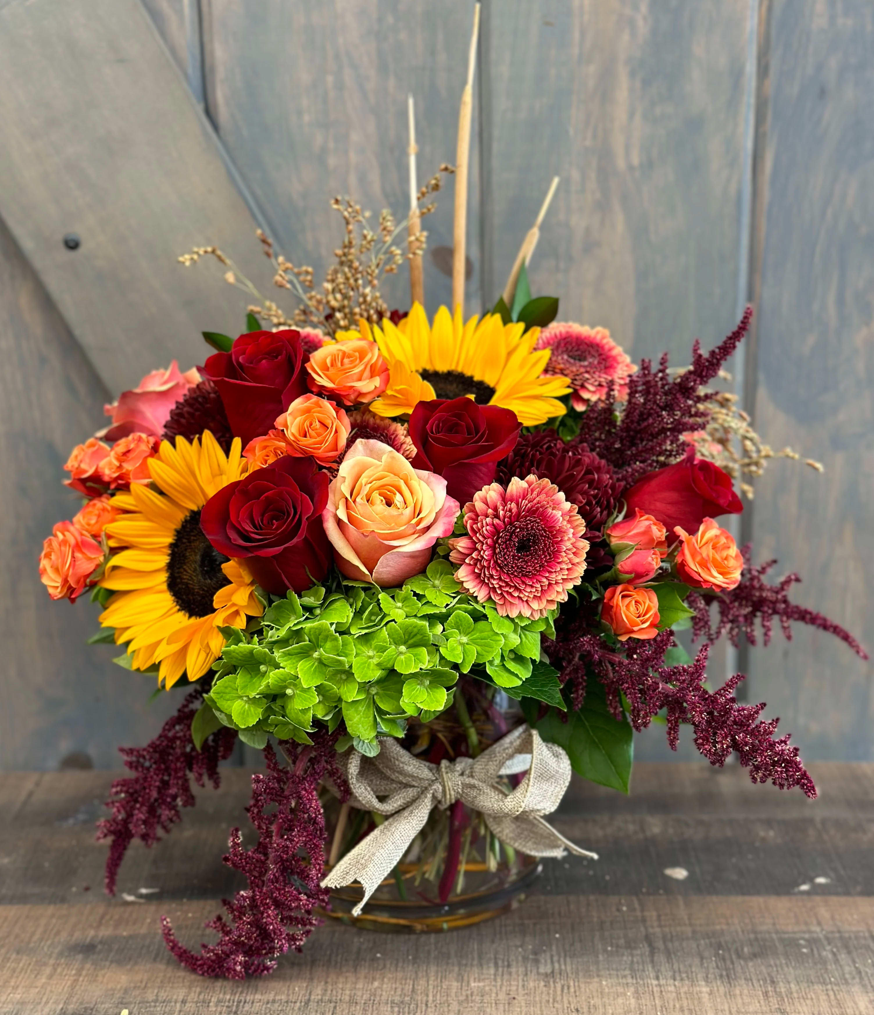 Fall in Love - This warm and vibrant mix is great for the loved one in your life this fall season. Included in this design are Red and Oranges Roses, Spray Roses, Sunflowers, Hydrangea, Chrysanthemums, Upright Amaranthus, gerbera Dasiys, Broom Corn, and Cattails