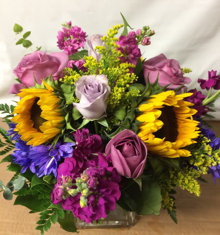 CAPE COTTAGE GARDEN' BOUQUET #CCGB - Cape Cod Cottage Vase A floral tribute to the charm of Cape Cod’s cottage gardens, this bouquet captures the essence of relaxed coastal elegance. Designed in the spirit of the classic Cape Cottage Garden—known for its informal, colorful blooms—this arrangement is perfect for any occasion or celebration.  Featuring bright sunflowers, berry-blush stock, lavender roses, and seasonal accents, it blends traditional cottage style with a fresh, modern twist. Arranged in a low vase, it’s ideal as a stunning table centerpiece or a cheerful room accent that brings the warmth and color of summer by the sea into any space. 