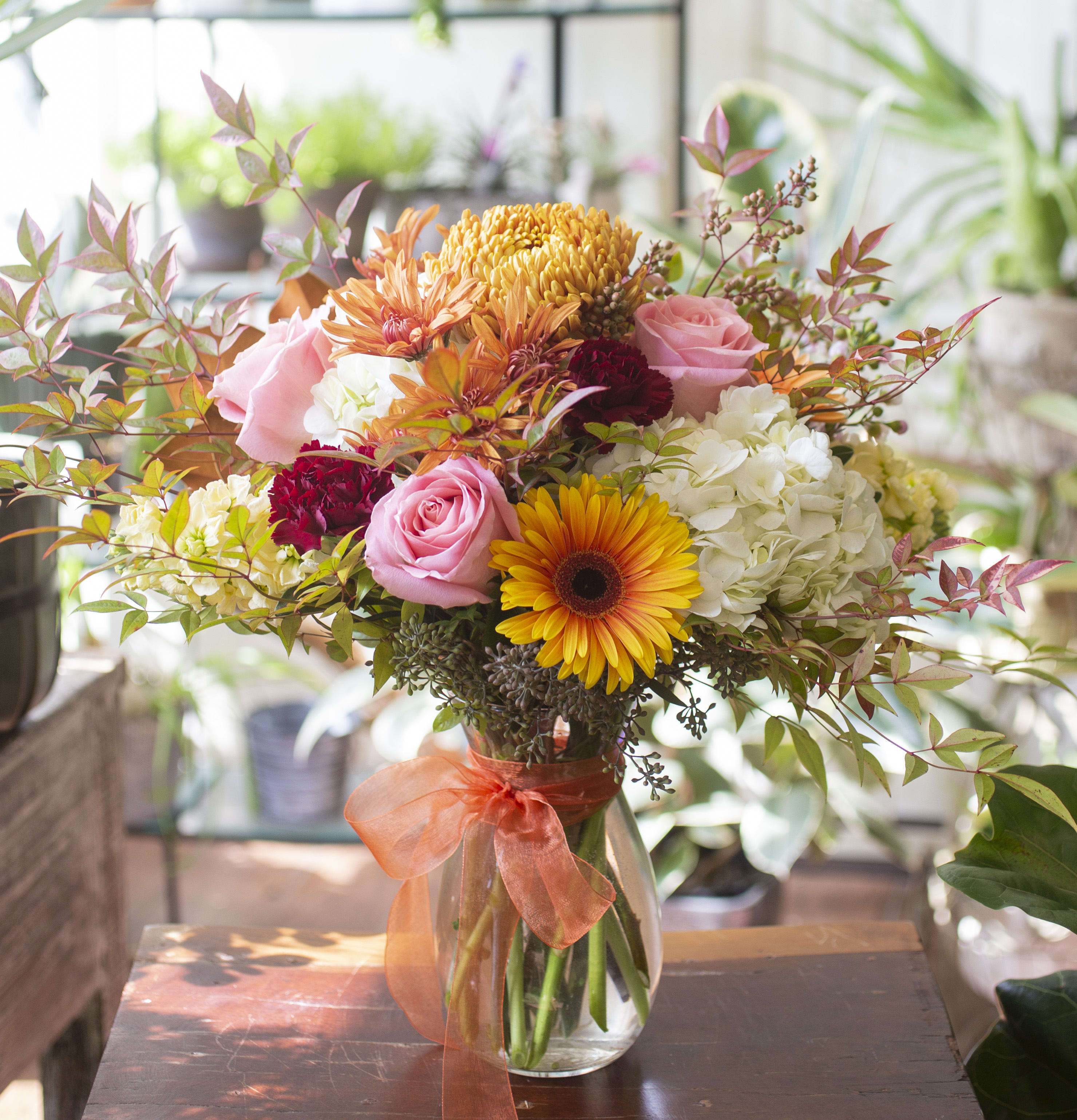 Paulette - Vibrant, warm and colorful blooms arranged in a clear glass vase with seasonal foliage and accents.