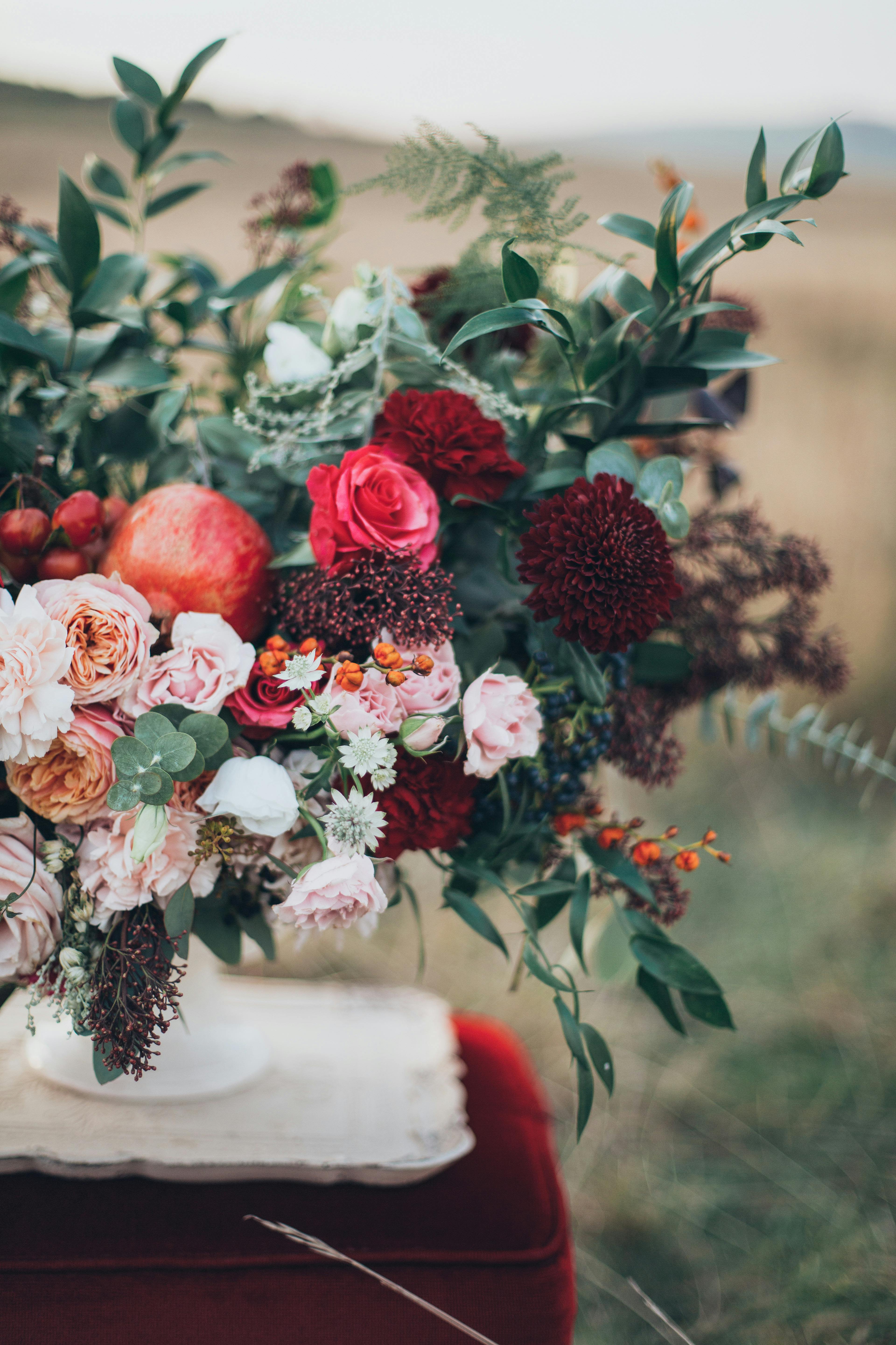 Lustre Noir - An opulent fusion of contrast and drama, this arrangement captures the essence of moody elegance. Velvety crimson dahlias, deep burgundy scabiosa, and rich red roses evoke mystery and sensuality, while creamy garden roses, pale lisianthus, and seasonal highlights offer a soft, luminous counterpoint. Accents of pomegranate, berry sprigs, and wild foliage add depth and texture, reminiscent of a Dutch Master’s still life. Both bold and refined, this composition whispers decadence and dark romance.