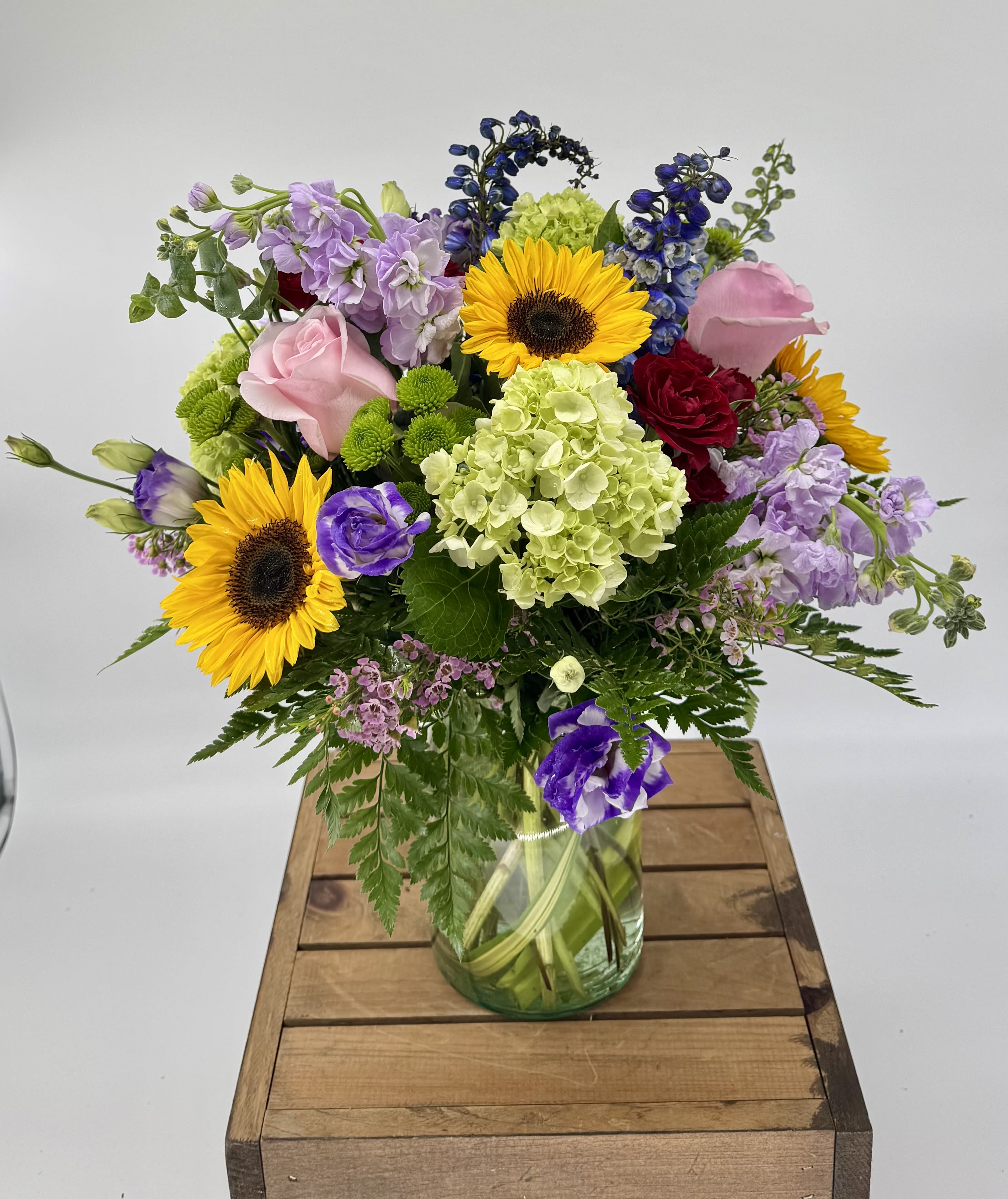 Colorful World (BR) - Brighten any space with this modern floral arrangement featuring vibrant sunflowers, green hydrangea, lavender stock, purple lisianthus, blue delphinium, red and pink roses, green buttons, and assorted greenery, beautifully arranged in a sleek clear glass vase. 