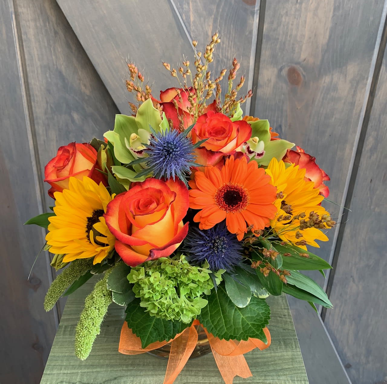 Autumn in the air - This tasteful centerpiece style vase arrangement is perfect to send during the autumn season. Our design includes Orange Roses, Mini green hydrangea, Green Cymbidium Orchids, Orange Alstromeria, Orange Gerbera Daisys, Yellow Sunflowers, Millet, and Blue Thistle all within a 5&quot; cylinder vase
