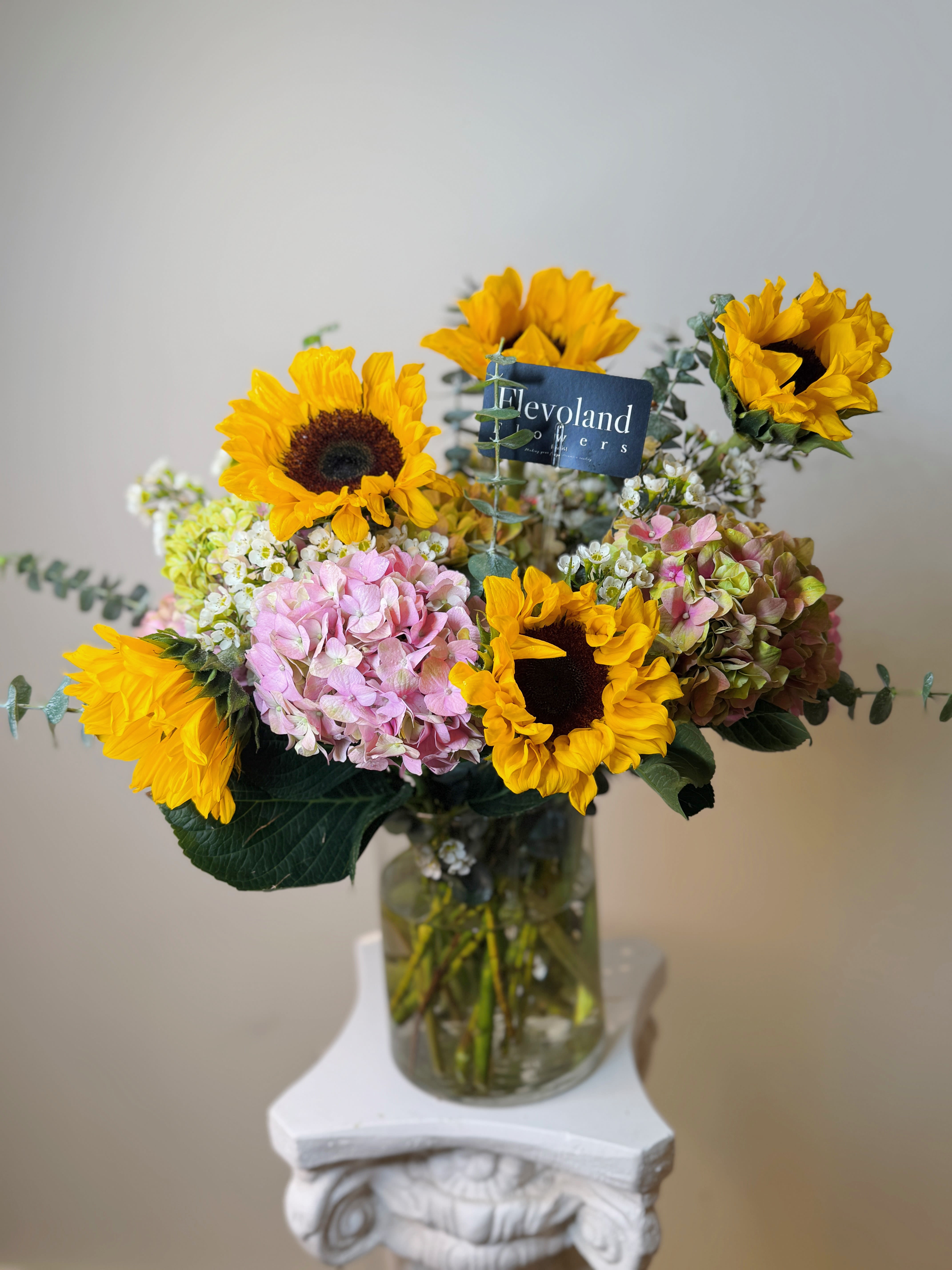 Sun Nostalgia - A bouquet that radiates both strength and elegance. Featuring 10 golden sunflowers, symbols of joy and vitality, paired with 10 lush hydrangeas that bring softness and refinement. Fresh eucalyptus stems add a touch of greenery and an uplifting fragrance, creating harmony between brightness and serenity.  Perfect for celebrating special moments, gifting heartfelt emotions, or simply bringing light into your home. Each design is crafted with care to leave a lasting impression.