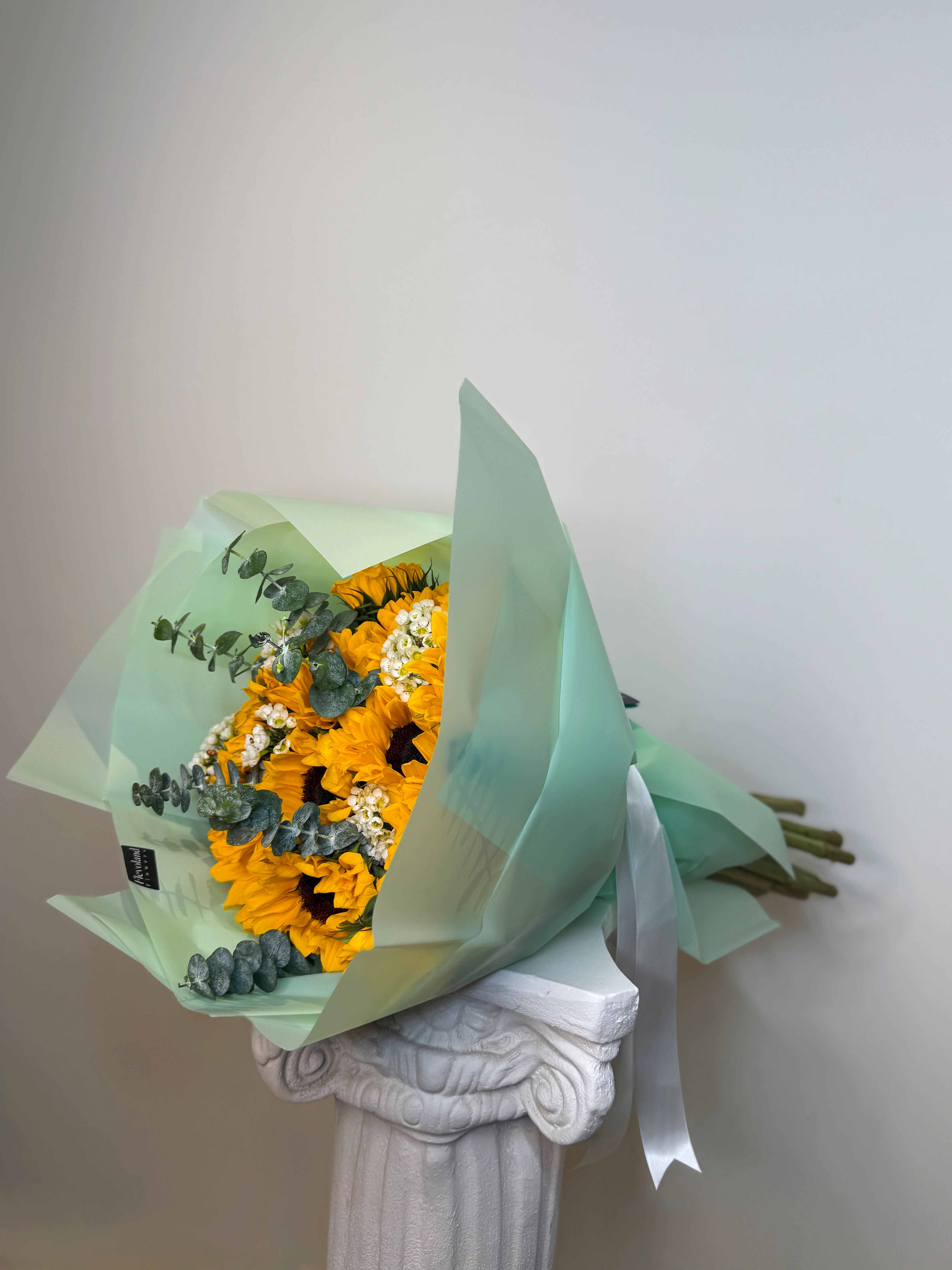 Clarté Solaire - A celebration of light and vitality. This bouquet of 20 sunflowers unfolds in full bloom, wrapped in soft green tones and highlighted with fresh eucalyptus and delicate accents. A design created to illuminate spaces with joy, elegance, and timeless beauty.