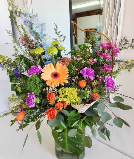 Hidden Garden Hidden Garden - A vibrant and eclectic garden-style arrangement bursting with color and texture. Featuring a cheerful peach gerbera daisy at the center, it’s surrounded by rich purple carnations, delicate blue delphiniums, and fiery orange spray roses and tulips. Accents of lime green button mums, soft green hydrangea, and whimsical greenery add volume and contrast. The mix of wildflowers, wispy fillers, and curly willow give it a free-spirited, organic feel—all beautifully gathered in a clear glass vase with lush eucalyptus foliage.