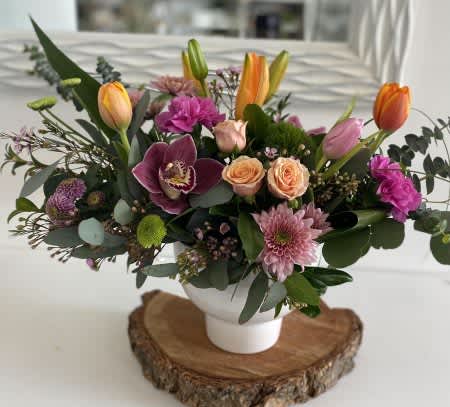 Valley Gardens - A stunning blend of natural charm and refined beauty, our Valley Gardens Centerpiece is a true showstopper. Nestled in a sleek white ceramic container, this arrangement features a lush combination of soft peach roses, exotic orchids, vibrant tulips, mauve chrysanthemums, and textured greens like eucalyptus and seeded eucalyptus.  Perfect for dining tables, gift-giving, or a thoughtful gesture of appreciation, this design balances wild garden textures with graceful blooms, making it ideal for modern rustic interiors or seasonal celebrations.