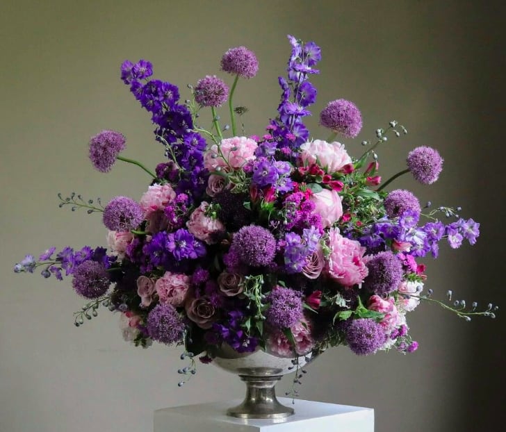 Violet Provance - A romantic burst of violets, blush, and lavender tones—this arrangement is a celebration of abundant texture and rich color. Featuring layers of allium, delphinium, roses, peonies, and heirloom florals, it is artfully gathered in a classic silver footed vessel. Grand and graceful, it evokes the opulence of a European salon or garden estate.  Perfect for elegant events, statement entryways, or an unforgettable gesture