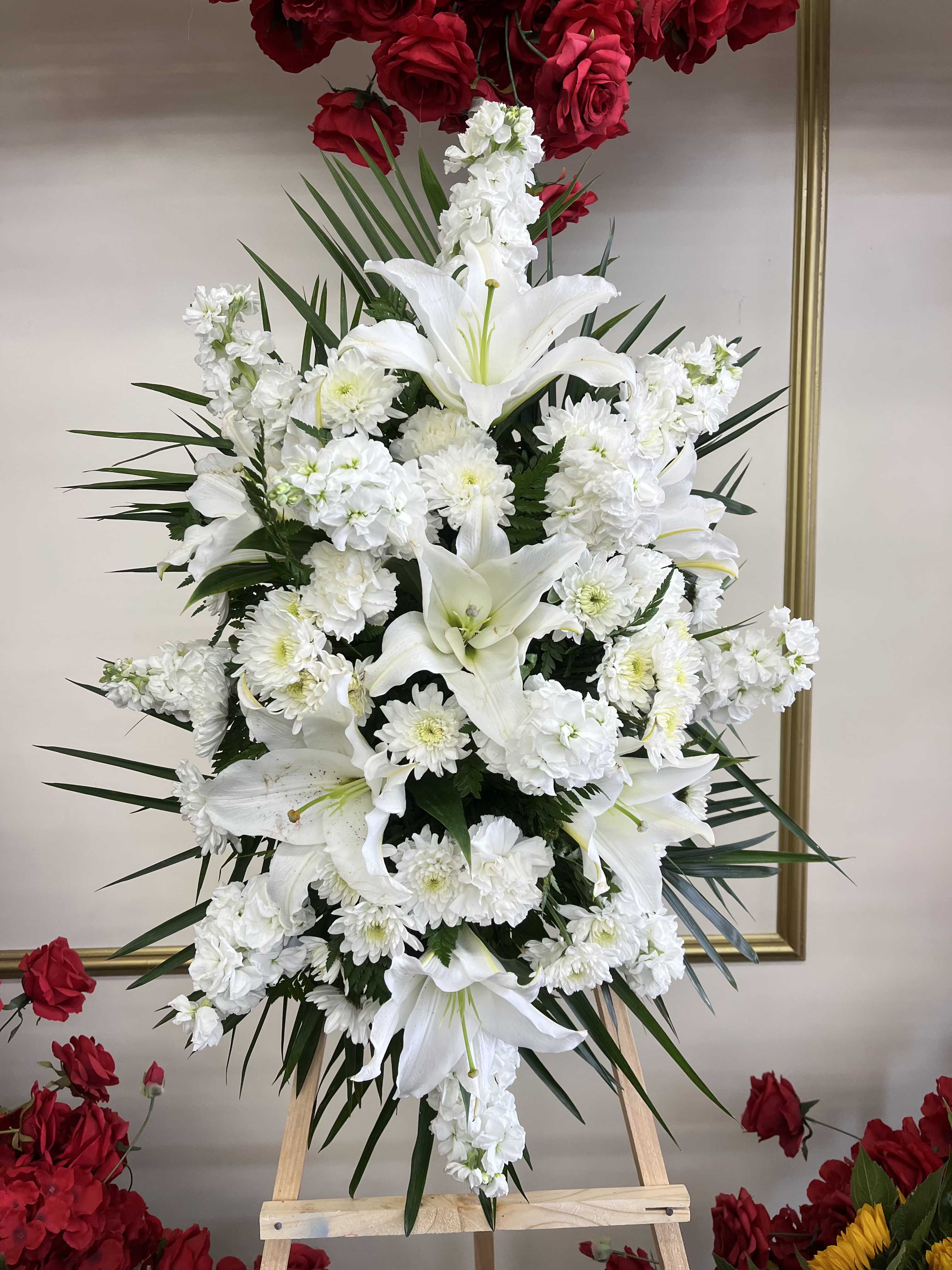 White Standing Spray - The White Standing Spray is an elegant display of sweet serenity. White roses, oriental lilies, stock, cushion pom and carnations are artfully arranged amongst emerald palm fronds and lush greens. Accented by white satin ribbon and standing on a wood easel this standing spray is an outstanding way to honor the life of your loved one.  Note: PICTURED IS THE STANDARD SIZE.  Easel is INCLUDED