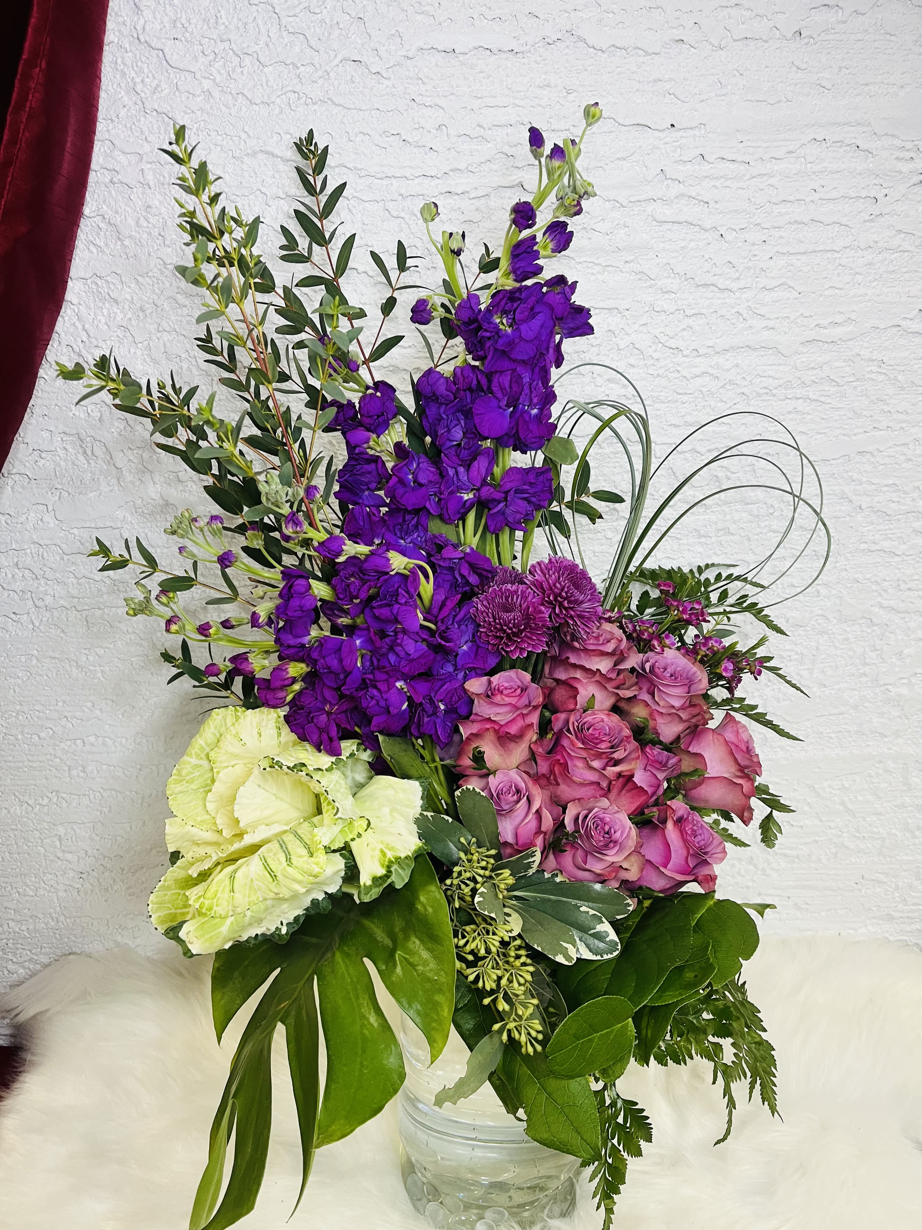 Amethyst Amethyst - Brighten any room with this vibrant and sophisticated flower arrangement, designed with a stunning mix of fresh, high-quality blooms. Perfect for special occasions, home decor, or as an unforgettable gift.   Features: 	•	Fresh flowers in rich tones of purple, lavender, and green 	•	Includes: Roses, stock flowers, chrysanthemums, ornamental cabbage, and a variety of lush greenery 	•	Artistic vertical design for a bold, modern touch 	•	Arranged in a clear glass vase with a textured pattern