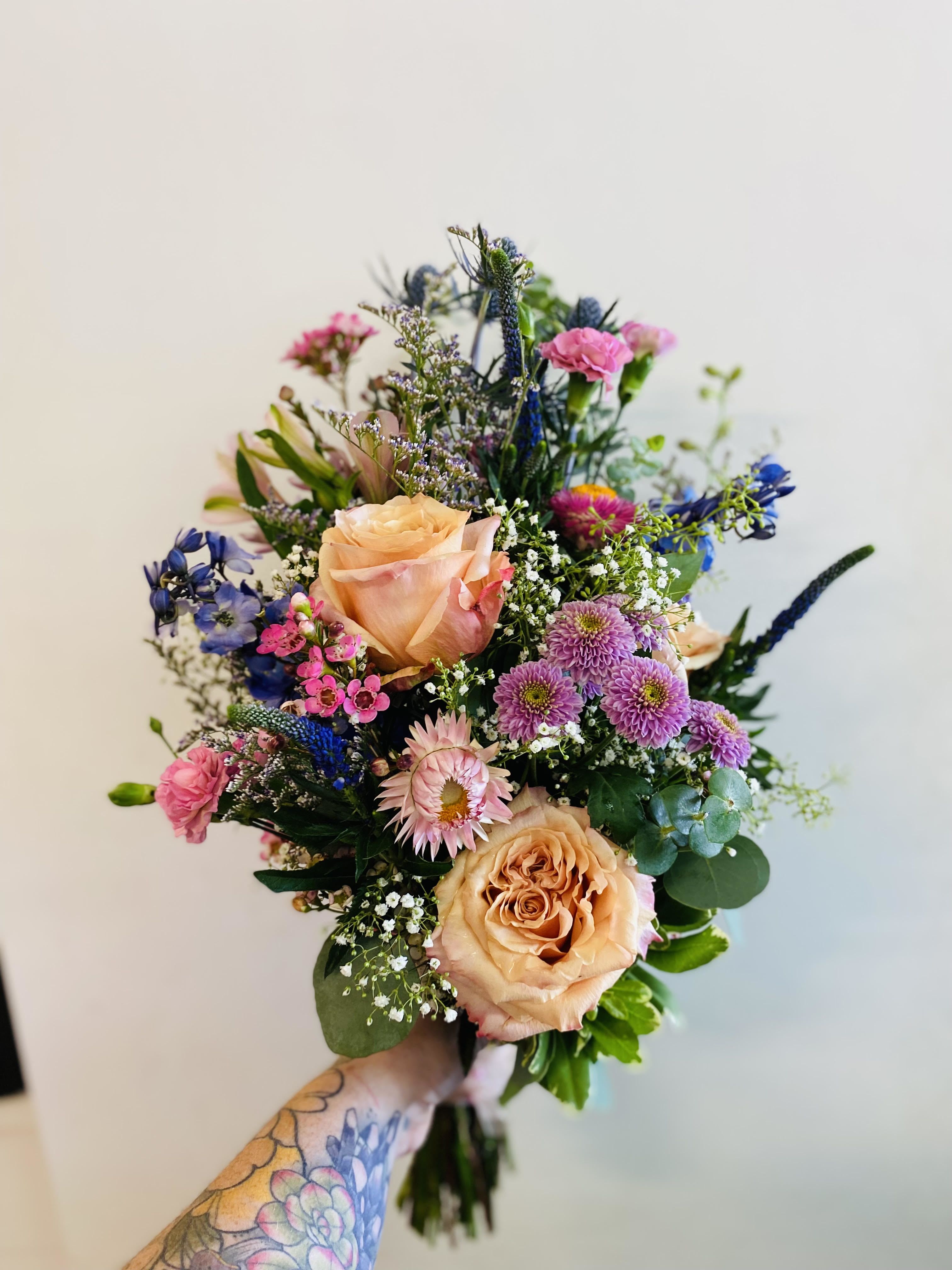 Petite Hand-Tied Bouquet Petite Hand-Tied Bouquet - This hand-tied bouquet is bursting with charm, blending warm peach roses with pops of vibrant pink, purple, and blue blooms. Textural accents like strawflowers, thistle, and baby's breath give it a wildflower-meadow feel, perfect for a romantic, garden-inspired look.