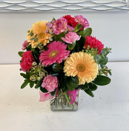 Rainbow Sherbet Rainbow Sherbet - This delightful bouquet features sunny peach and pink gerbera daisies, vibrant carnations, all nestled in lush greenery. Arranged in a clear square glass vase with a playful pink ribbon, it’s a joyful display perfect for celebrations or simply spreading smiles.