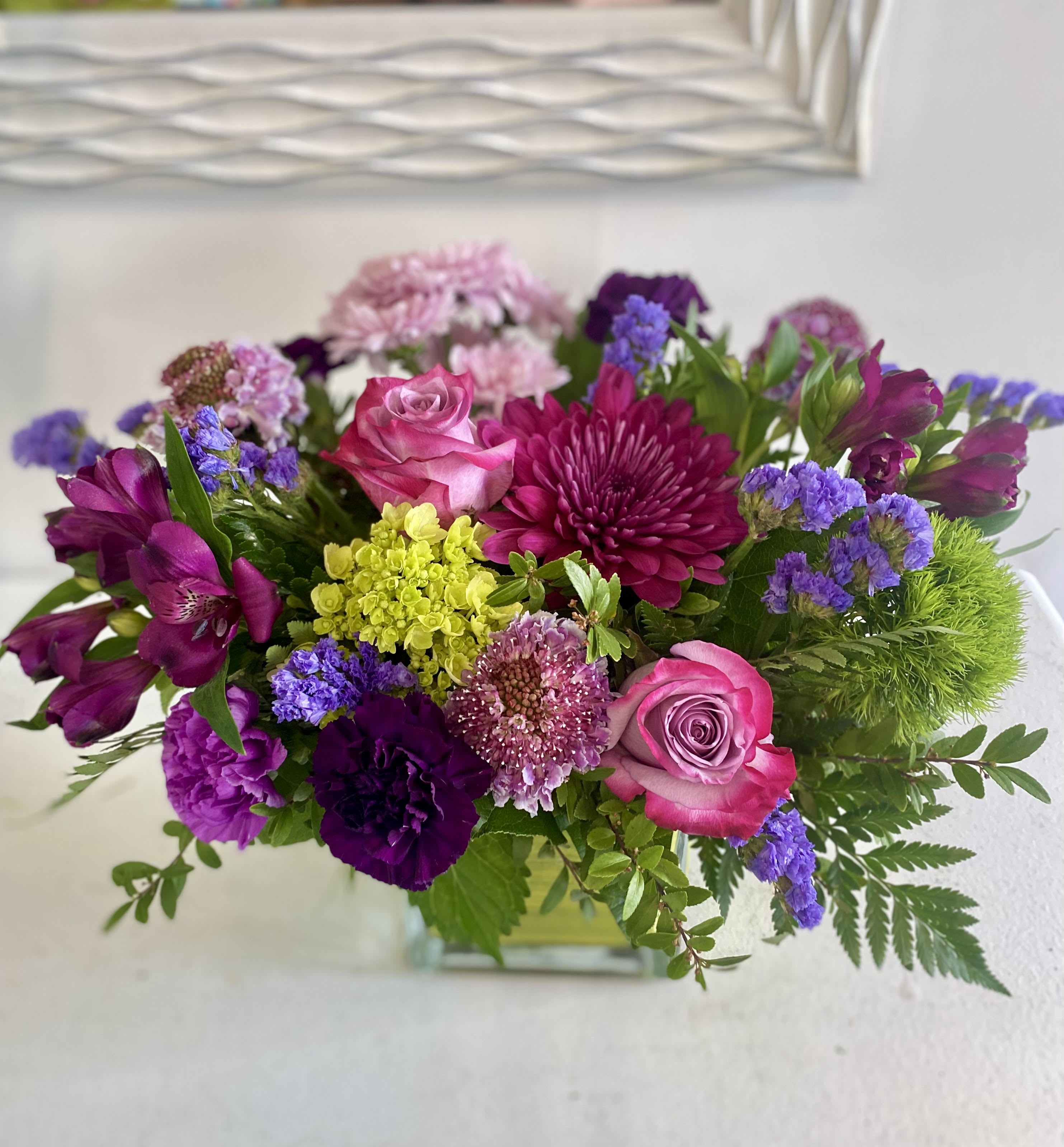 Purple Passion - A vibrant mix of magenta, purple, and soft pink blooms creates a rich, eye-catching display. Featuring roses, chrysanthemums, carnations, and scabiosa, accented with pops of yellow and textured greenery, all arranged in a clear square glass vase. This arrangement is bold, cheerful, and full of personality.