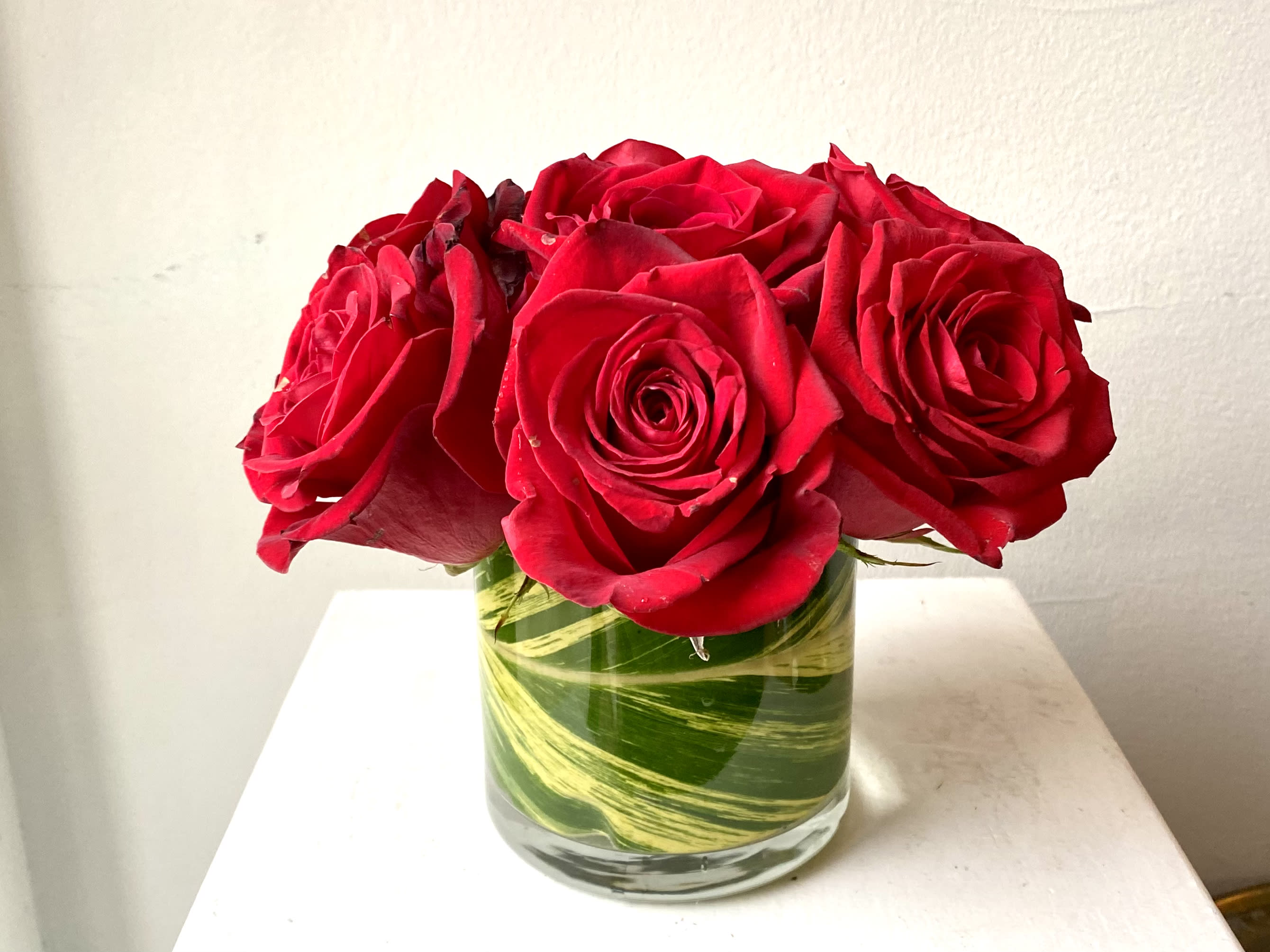 Simple Elegance Simple Elegance - 5 Red Roses in a low round glass vase enveloped in a tropical leaf.