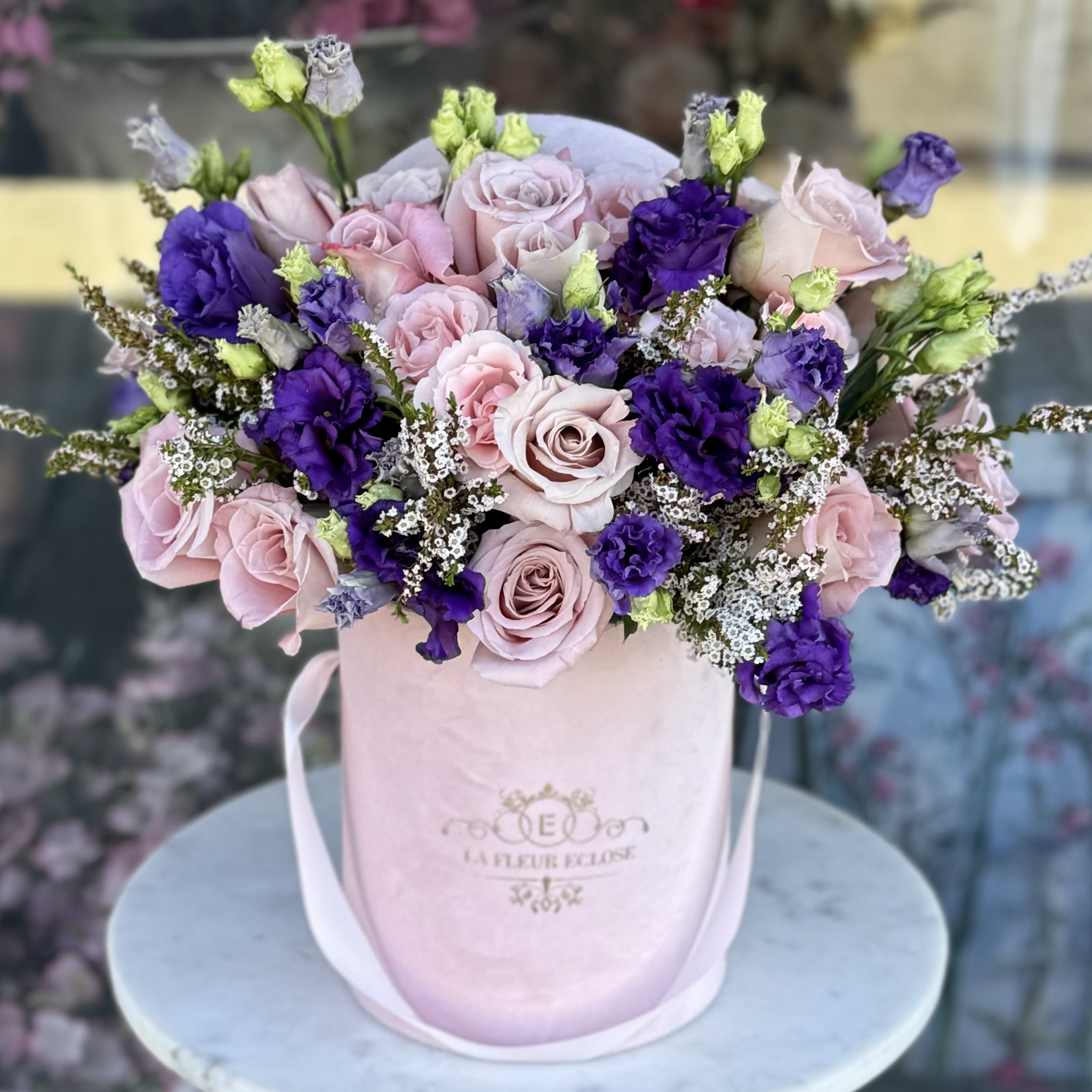 Product 13 Product 13 -  A stunning mix of soft pink roses, lush purple lisianthus, and delicate white waxflowers. The pastel tones blend seamlessly with vibrant purple hues, creating a charming and sophisticated bouquet. Hints of fresh green lisianthus buds add a lively touch, making this arrangement truly captivating.  Presented in a chic velvet hatbox, this design is perfect for expressing love, celebrating special occasions, or adding a touch of luxury to your space. Whether as a gift or a centerpiece, Elegant Romance is sure to leave a lasting impression.