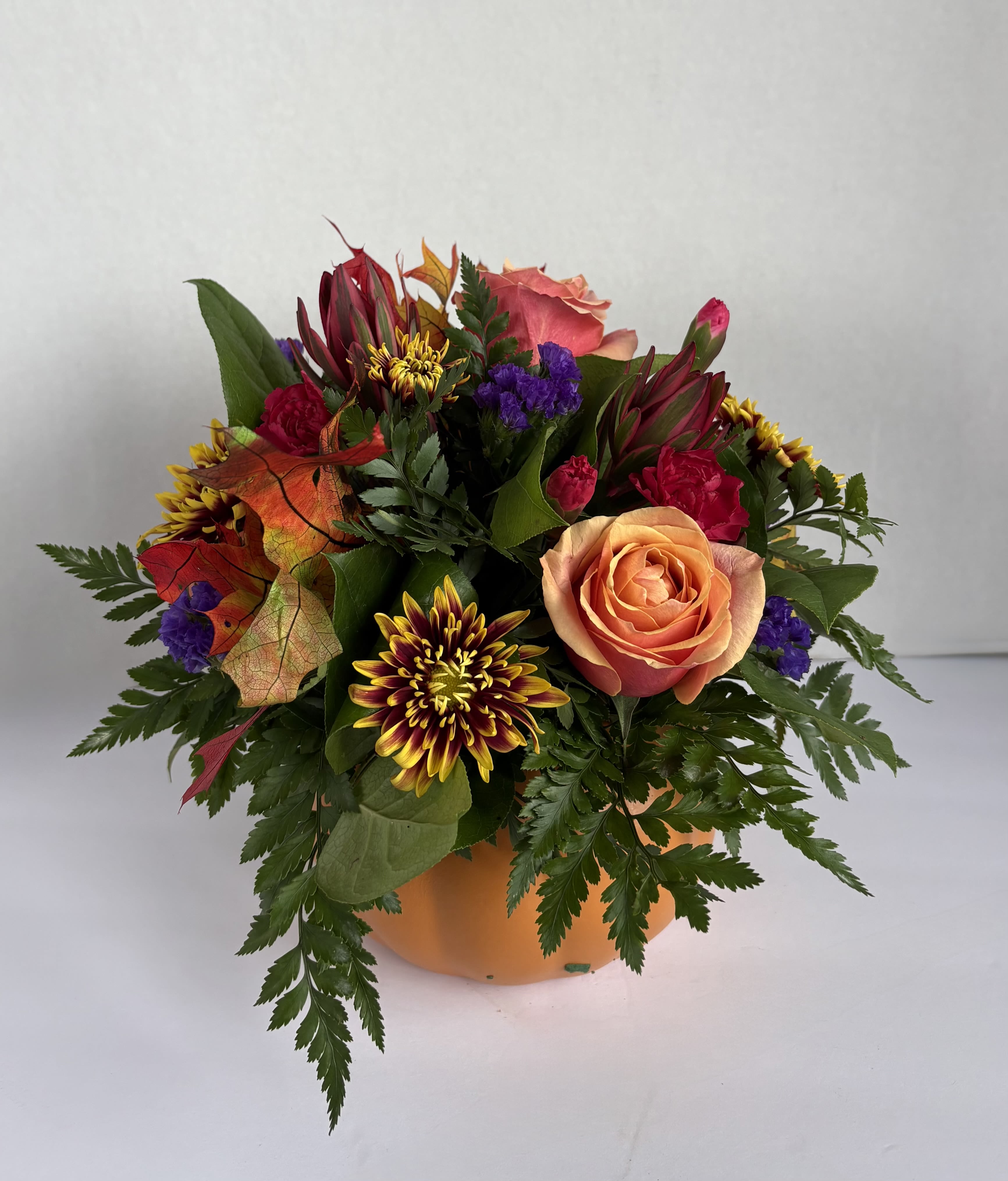 Fall is in the Air - Beautiful Fall arrangement with orange roses, purple statice, mums and leave and greens in orange pumpkin shaped container.