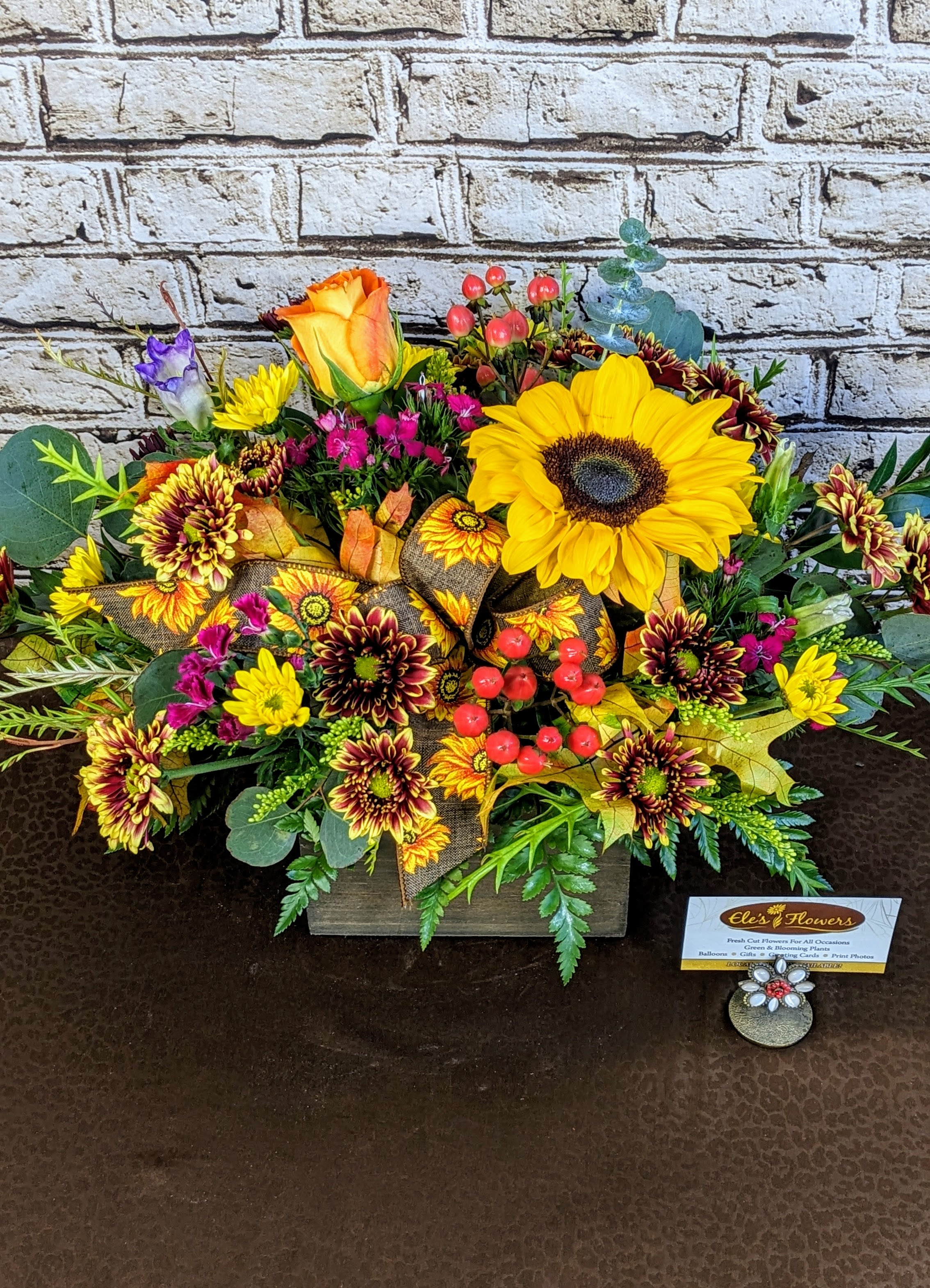 Fall Blooms Box - This fall bloom arrangement in a rustic wooden box brings the rich colors of the season indoors. Featuring a mix of warm-toned flowers like sunflowers, mums, and more, it’s accented with autumnal foliage, dried grasses, and seasonal elements like berries or wheat. The wooden box adds a natural, farmhouse charm, making this arrangement a perfect centerpiece or decor piece that captures the cozy, earthy essence of fall.  The designs we create are custom and the blooms and containers will vary. We use the freshest product available to create the perfect floral combination for the recipient. If you have any special requests, please add those in the &quot;special instructions&quot; portion of your order to help us dial in your vision. Otherwise, our skilled designers, Sara &amp; Brett, will take it from here!  *Please note that the photos are examples only – floral selection will vary.*  Standard Size is a great centerpiece size (thinking table size) Approx. Dimensions:16”H x 12”W 