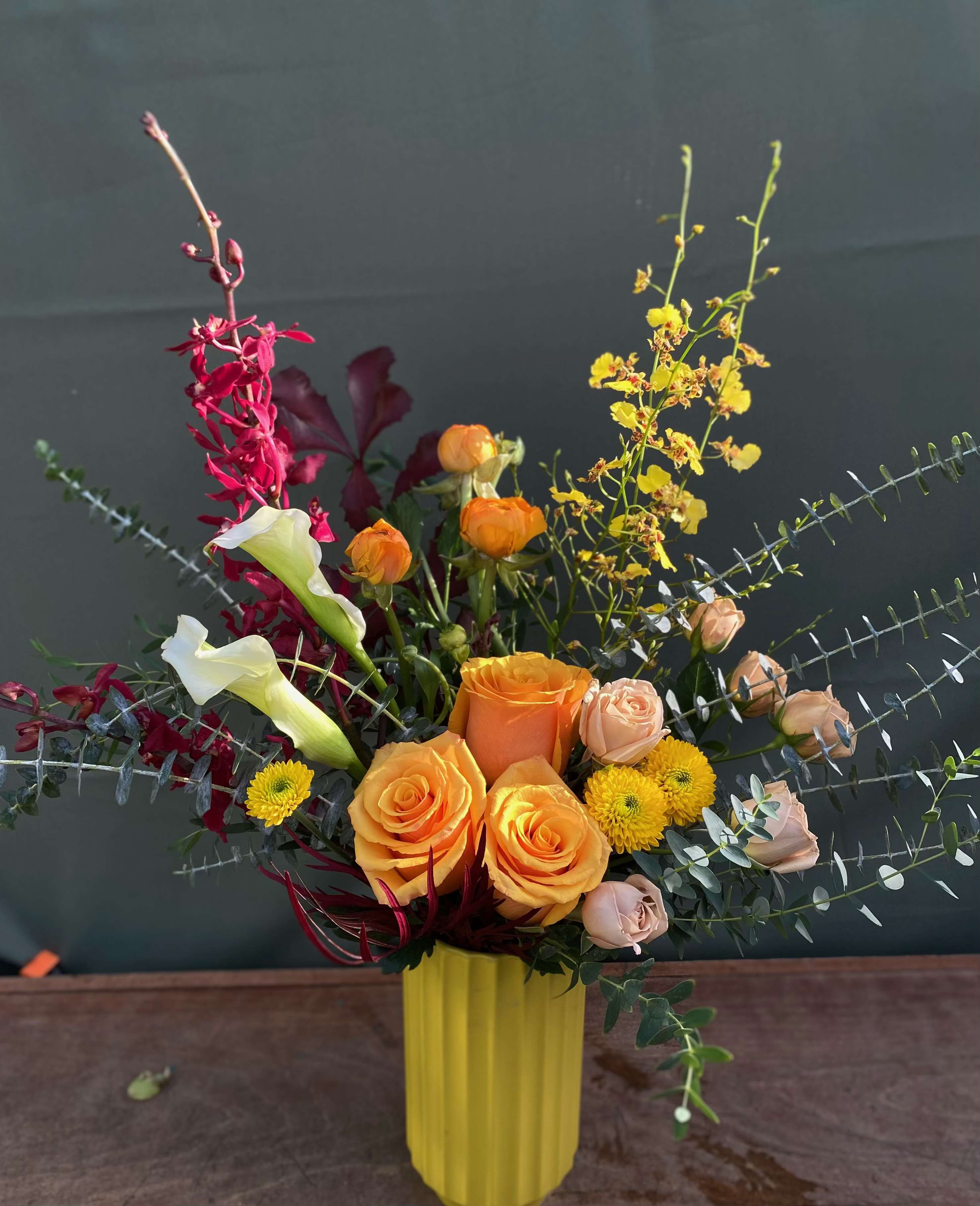 Gleeful - This arrangement includes orange roses, white calla lilies, eucalyptus, orchids, ranunculus all hand arranged in a cheerful yellow ceramic vase.