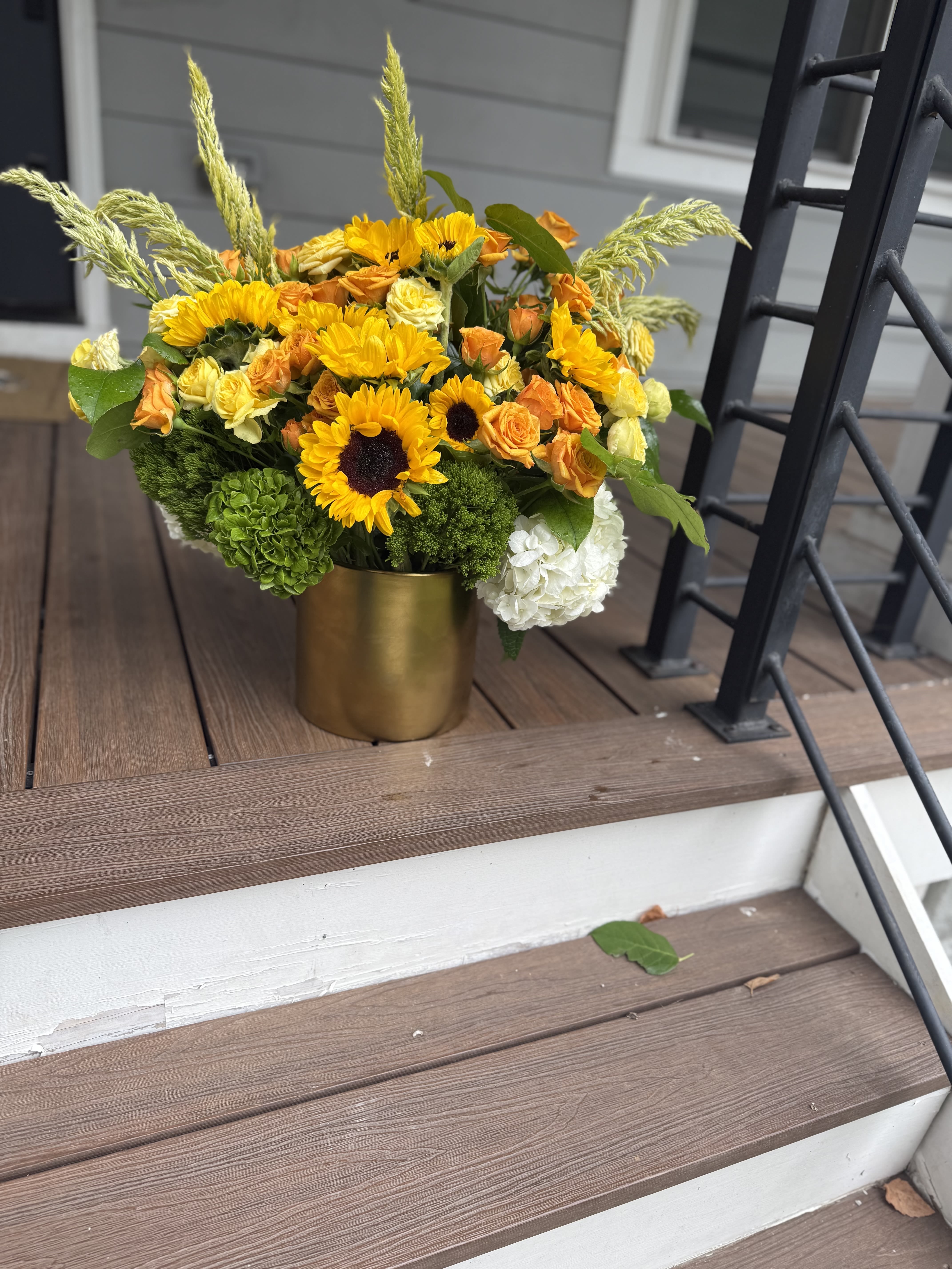 The Golden Child - Radiant, bold, and full of joy — our Golden Child bouquet is a celebration of sunshine and abundance. Featuring vibrant sunflowers, golden spray roses, lush hydrangeas, and rich greenery, this arrangement is designed to brighten any space and lift every spirit. The golden tones symbolize warmth, gratitude, and new beginnings, while the textured greens add depth and balance. Presented in a sleek metallic vase, Golden Child is perfect for celebrating milestones, gifting to someone special, or simply bringing light into your home