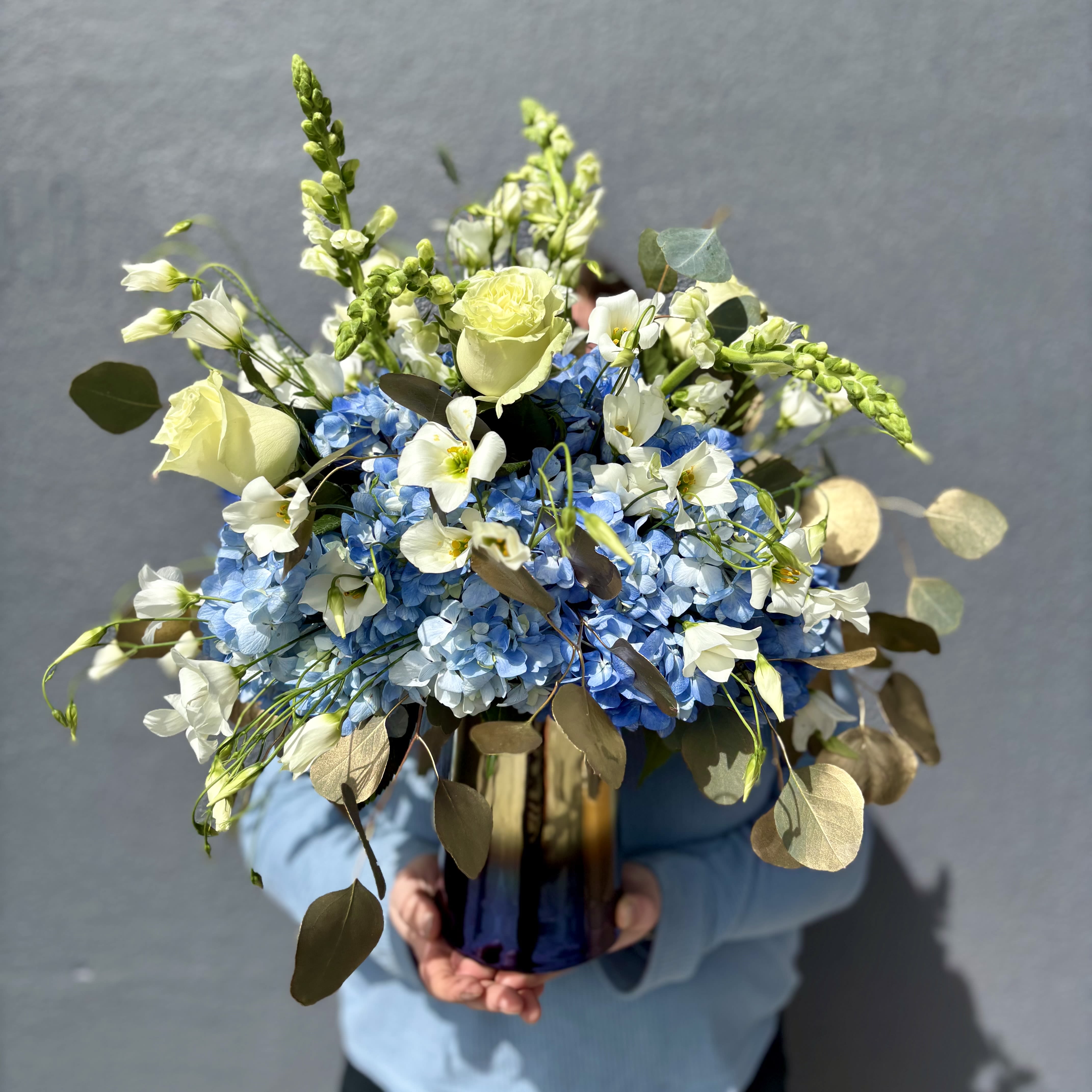 Heart of Gold - Heart of Gold is an arrangement that will really shine. Fluffy blue hydrangeas sit as a base for a ring of white snapdragons and roses and sprinkled with lisianthus. This design is then taken to the next level with gold dusted silver dollar eucalyptus. All neatly arranged in a gold and blue vase. 