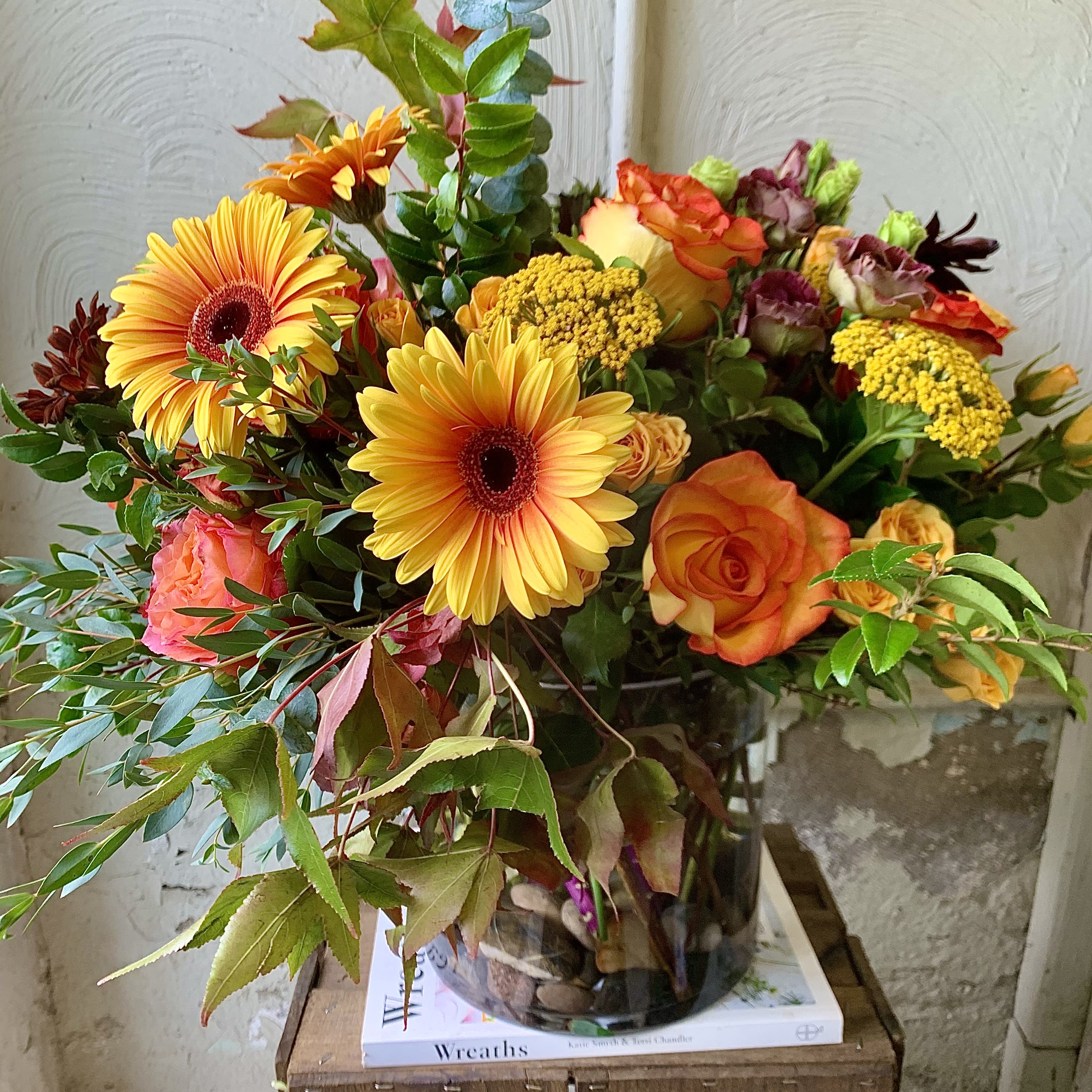 October Skies - A natural and rustic mix of garden-fresh varieties showcasing gerbera, orange roses and seasonal fillers, designed in a chic glass vase with lush greens and berries.