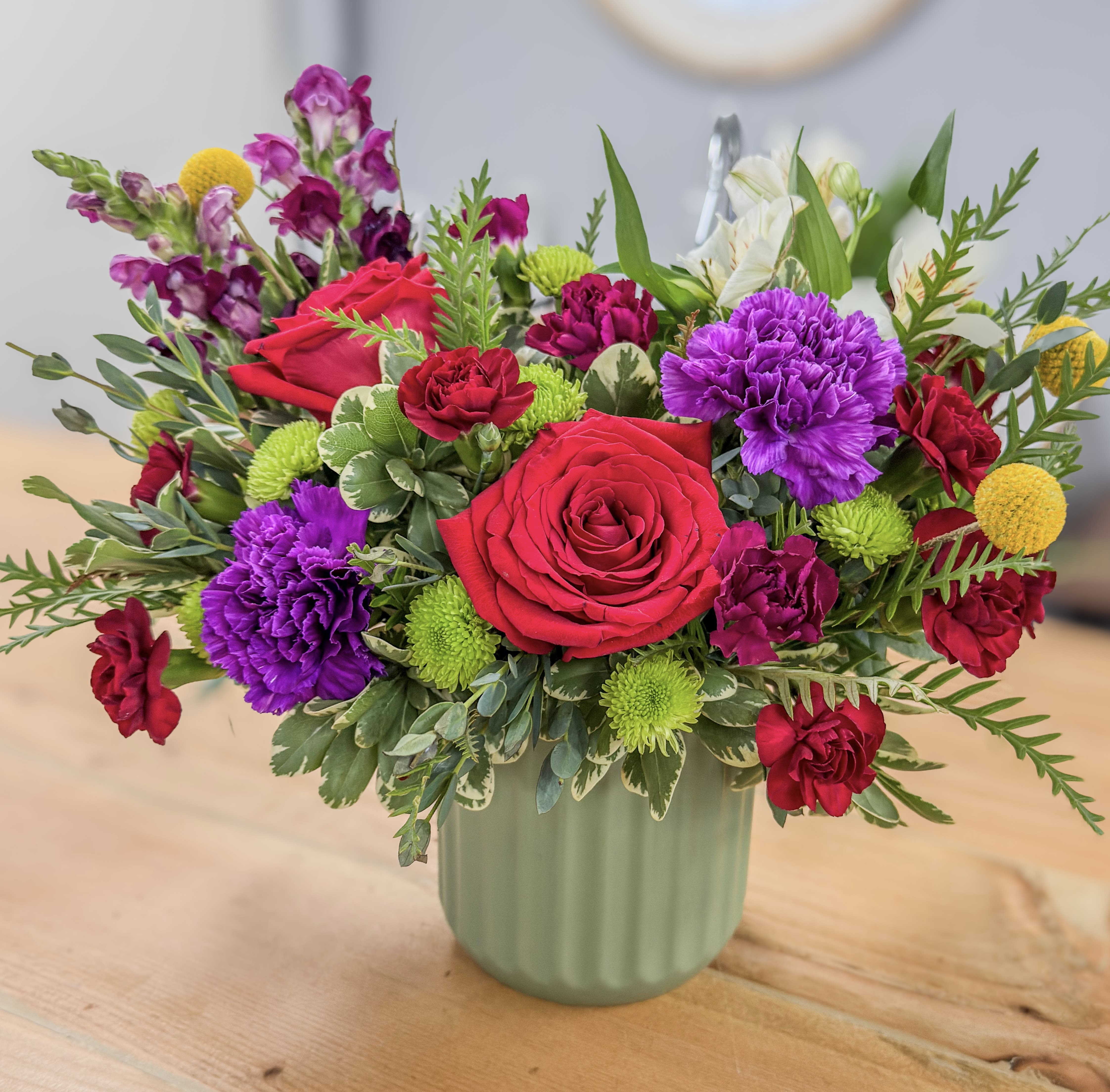 The Jewel Tones Bouquet - A stunning and vibrant arrangement that's perfect for any occasion. This beautiful bouquet features a bold mix of deep red roses, rich purple and red carnations, and striking green blooms. Accented with pops of yellow, this colorful mix is beautifully presented in a modern, sage green vase.