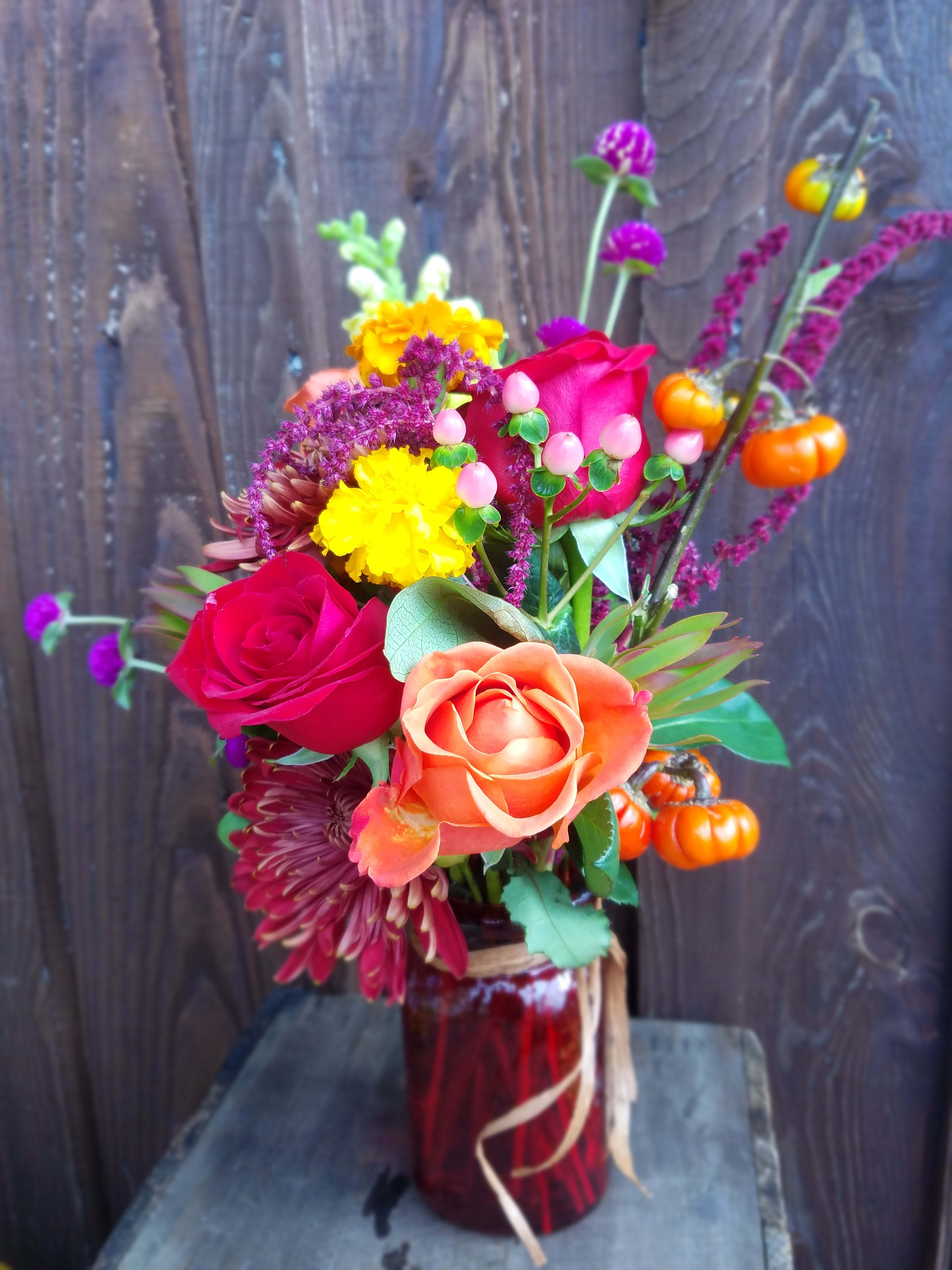 Harvest Bounty - This deep colored bouquet of fresh flowers will bring the rich colors of harvest season to your home featuring roses, mums, amaranth, gomphrena, marigolds and hypericum. It even features our own home-grown 'pumpkin on a stick'!  These flowers are overflowing from a deep ruby red hammered glass jar
