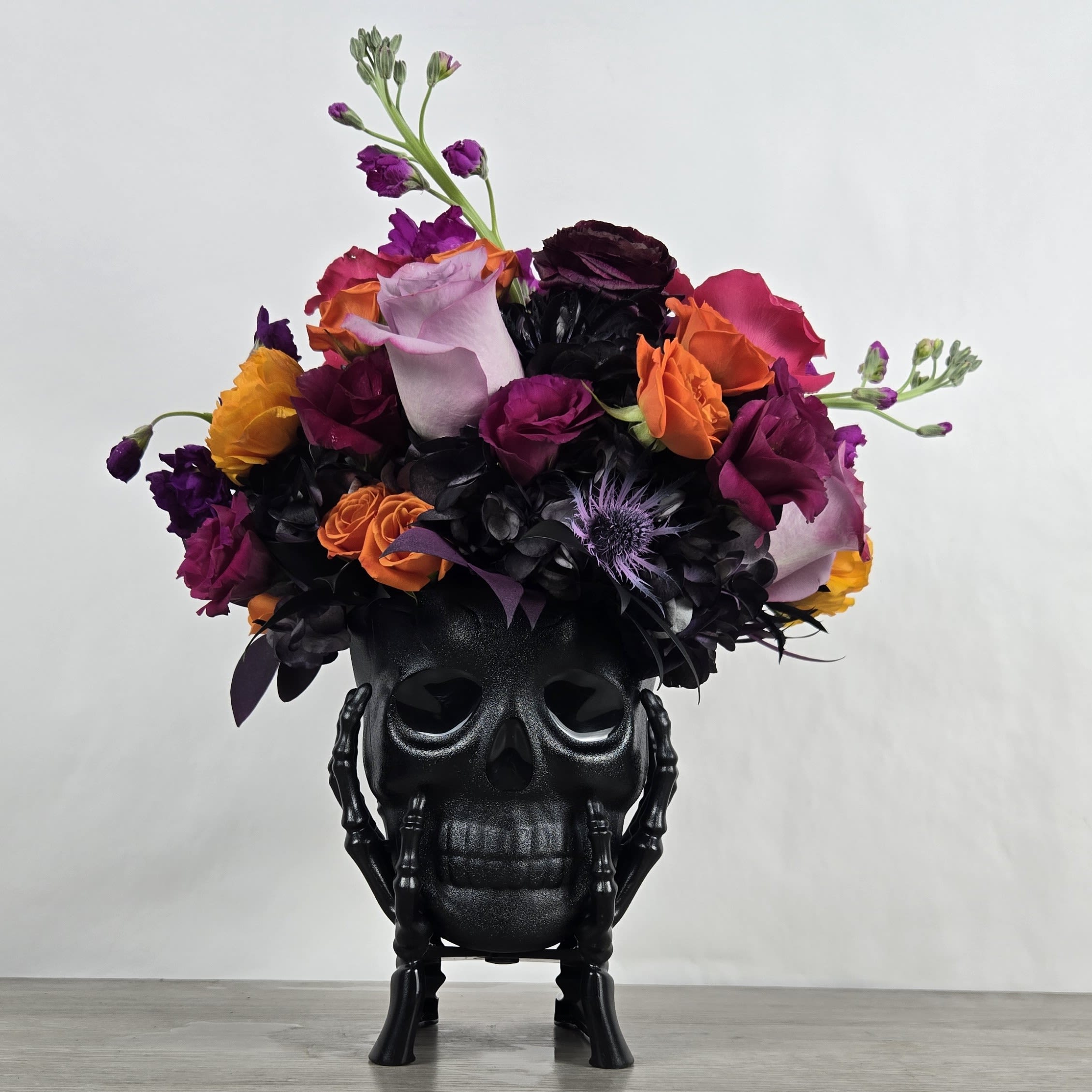 Calavera - Calavera is a bold and bewitching Halloween floral arrangement inspired by the vibrant spirit of Día de los Muertos. This dramatic display blends deep purples, fiery oranges, sunlit yellows, and inky blacks with a whisper of pink — a nod to both the mystery of the season and the beauty of remembrance.