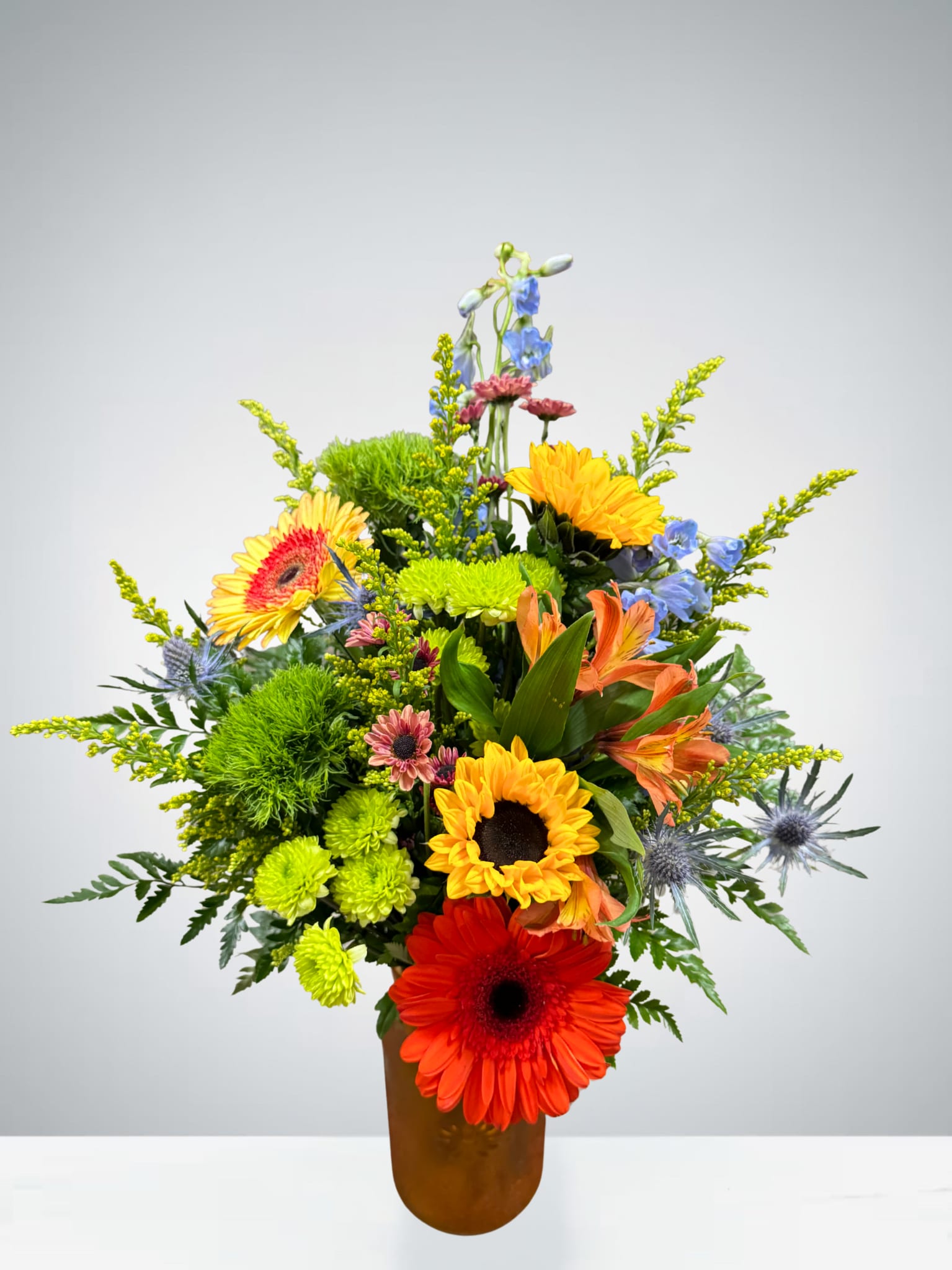 Joyful Moments  - A cheerful burst of color, this arrangement features vibrant sunflowers, gerbers, daisies, blue delphiniumn, alstroemeria, chrysanthemums, and more, accented with fresh greenery and pops of blue thistle. Designed in a frosted vase, it’s perfect for brightening someone’s day or celebrating life’s happy moments.